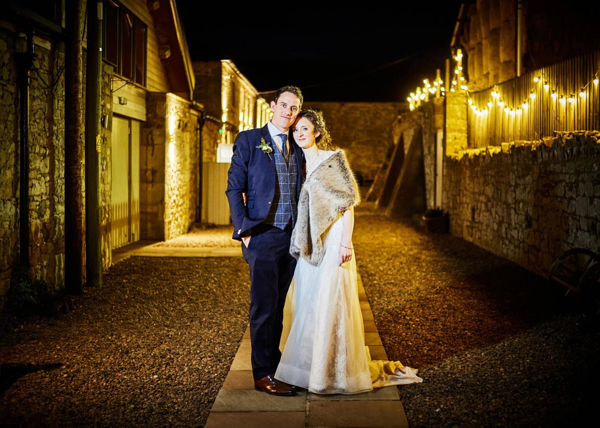 A couple dressed in wedding attire standing close together outside at night. The man is in a navy suit with a white shirt and plaid vest, and the woman is in a white wedding dress with a fur shawl. They are on a small wooden walkway with buildings an