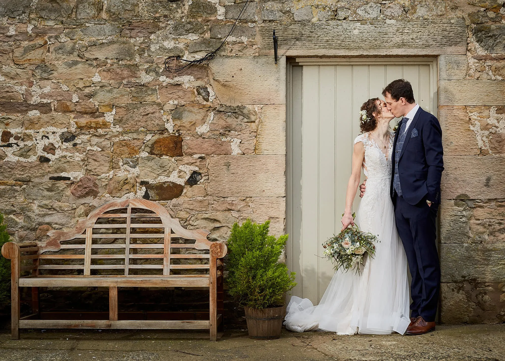 A bride and groom sharing a kiss outdoors, standing in front of a rustic stone wall with a door, a wooden bench, and potted plant nearby.