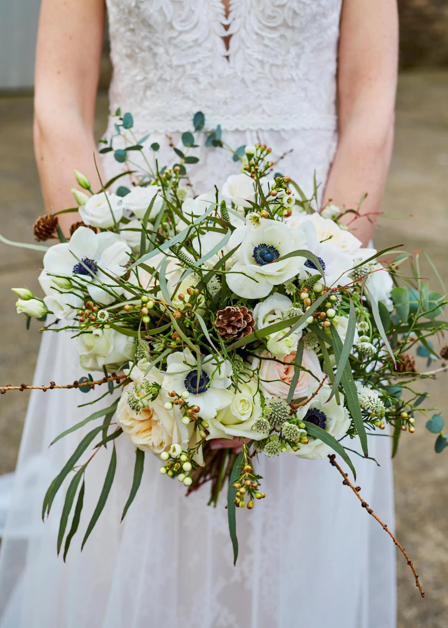 A bride holding a bouquet of white flowers, greenery, and small pinecones, wearing a white dress with lace details.