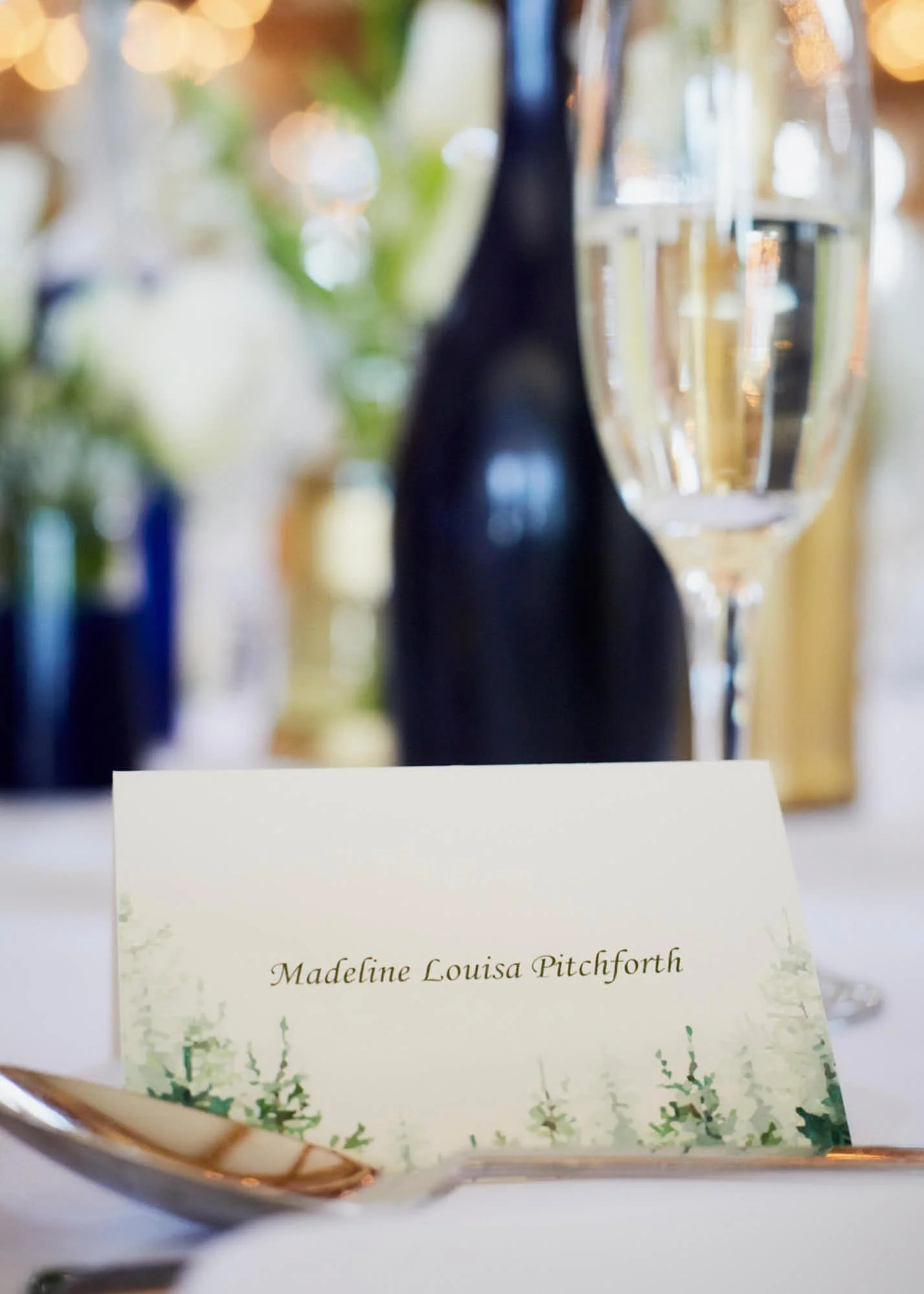 Name tag with the name Madeline Louisa Pitchforth in elegant script in front of a blurry background of glassware and lights.