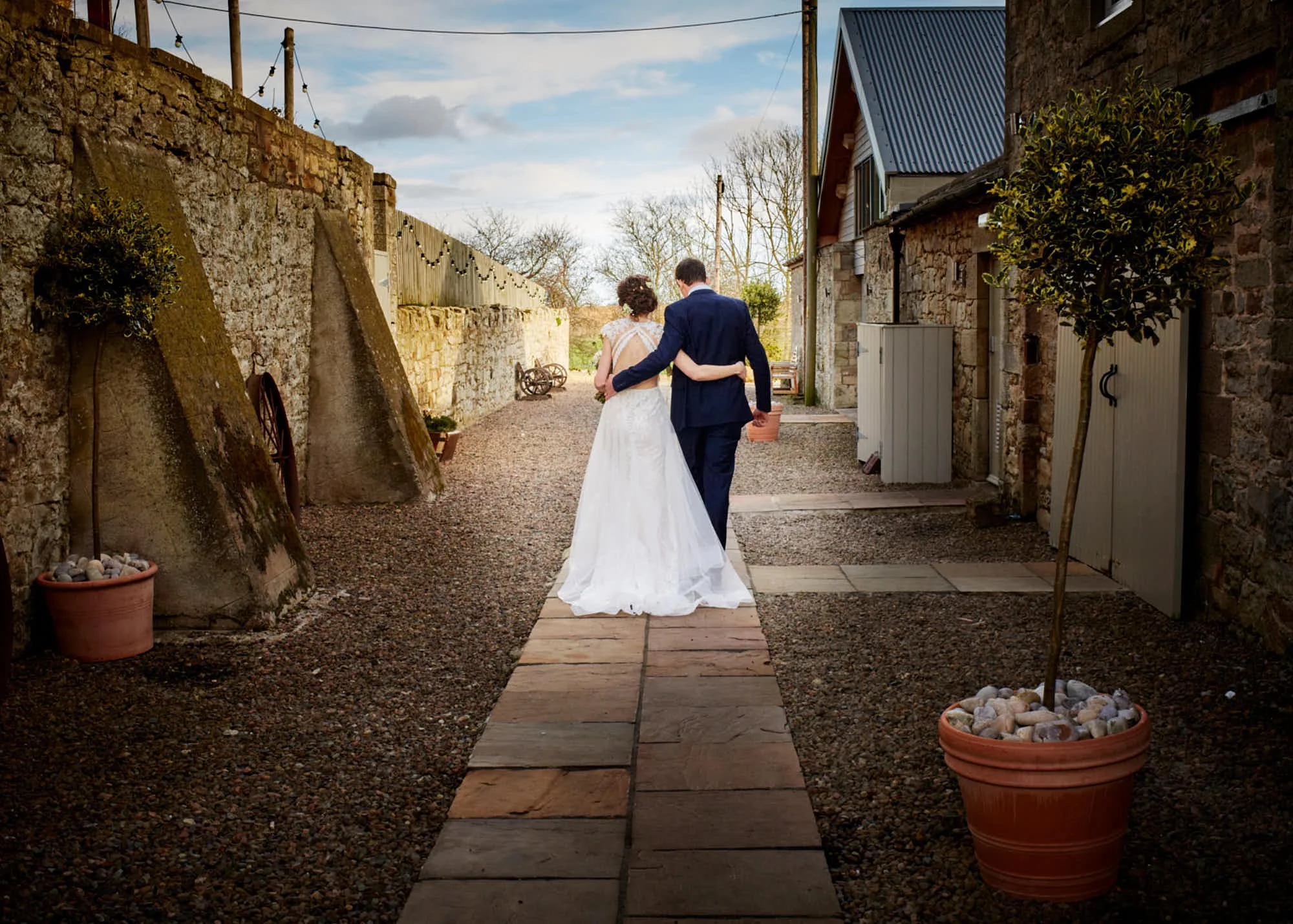 A bride and groom walking arm in arm along a stone path outside a rustic stone building. The bride is wearing a white wedding dress, and the groom is in a navy blue suit. The scene is set during the day with a partly cloudy sky, and there are potted 