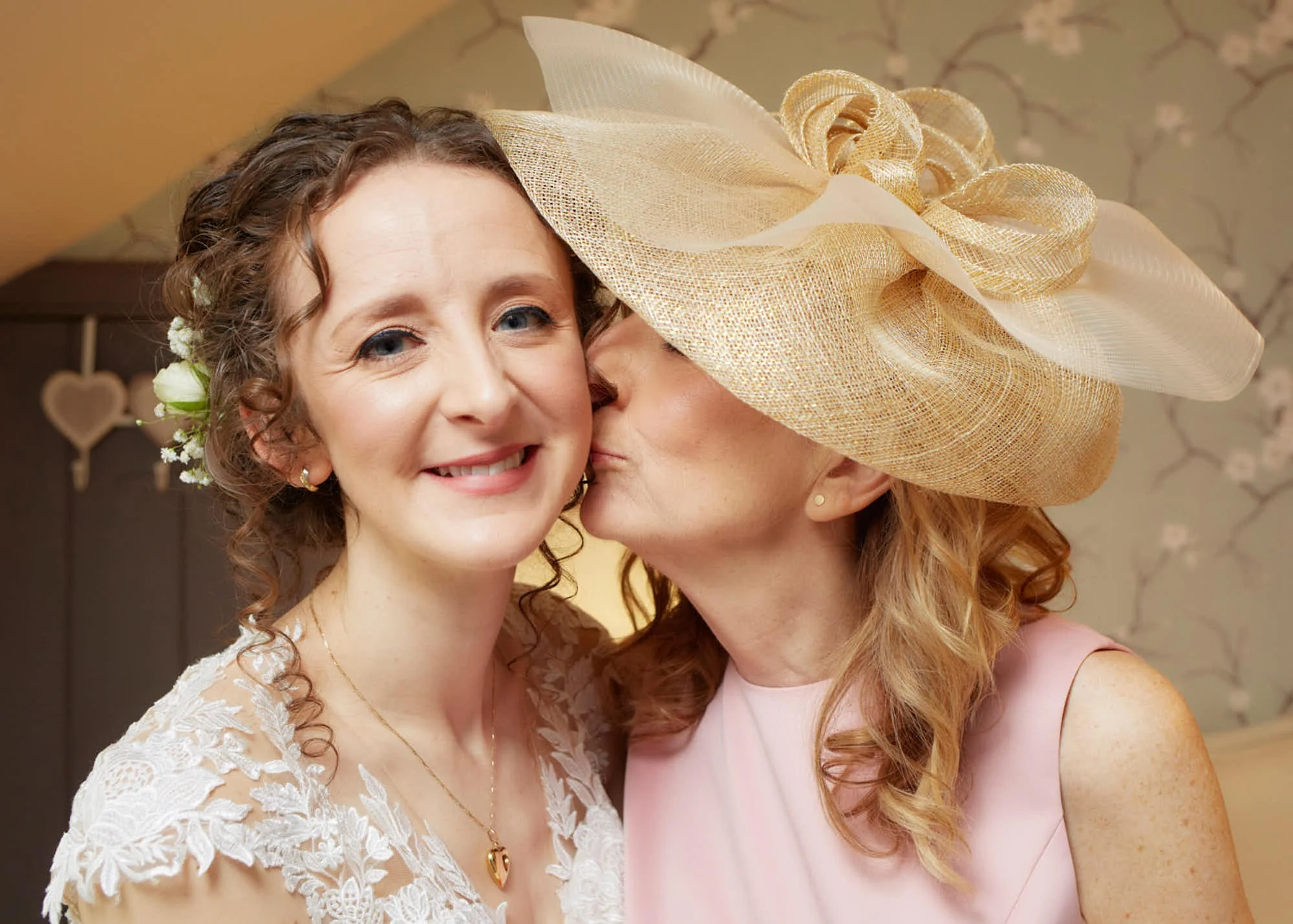 Two women celebrating a special occasion, with one woman kissing the other on the cheek. The woman on the left has curly hair, is smiling, and is wearing a lace dress with a floral headpiece. The woman on the right has red hair, is wearing a large be