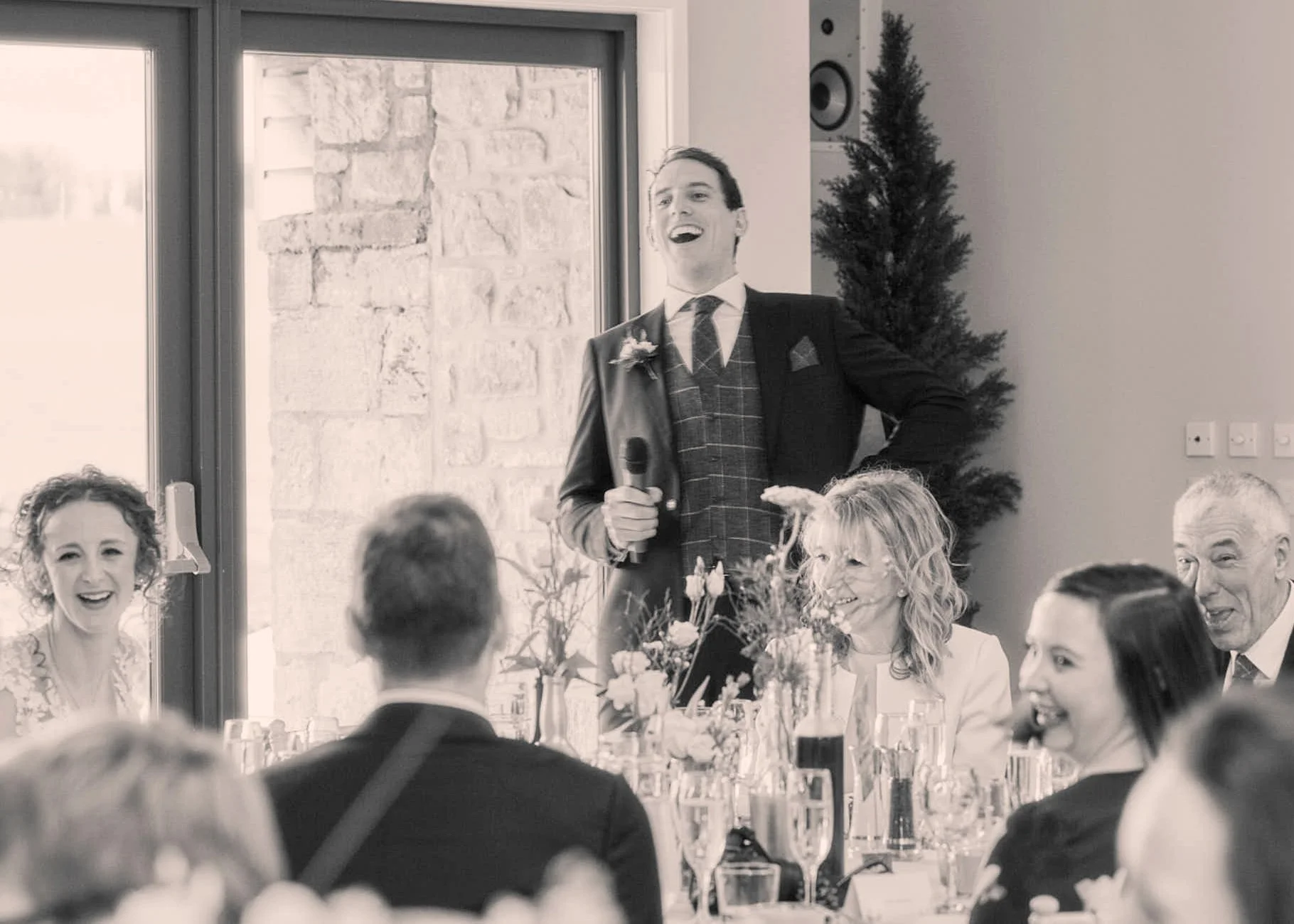 A man in a suit with a microphone giving a speech at a wedding reception, surrounded by seated guests at decorated tables.