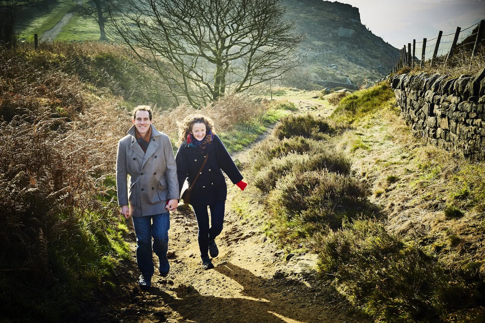 A young man and woman walking on a dirt trail in a rural, hilly landscape with shrubs, trees, a stone wall, and hills in the background during daytime.