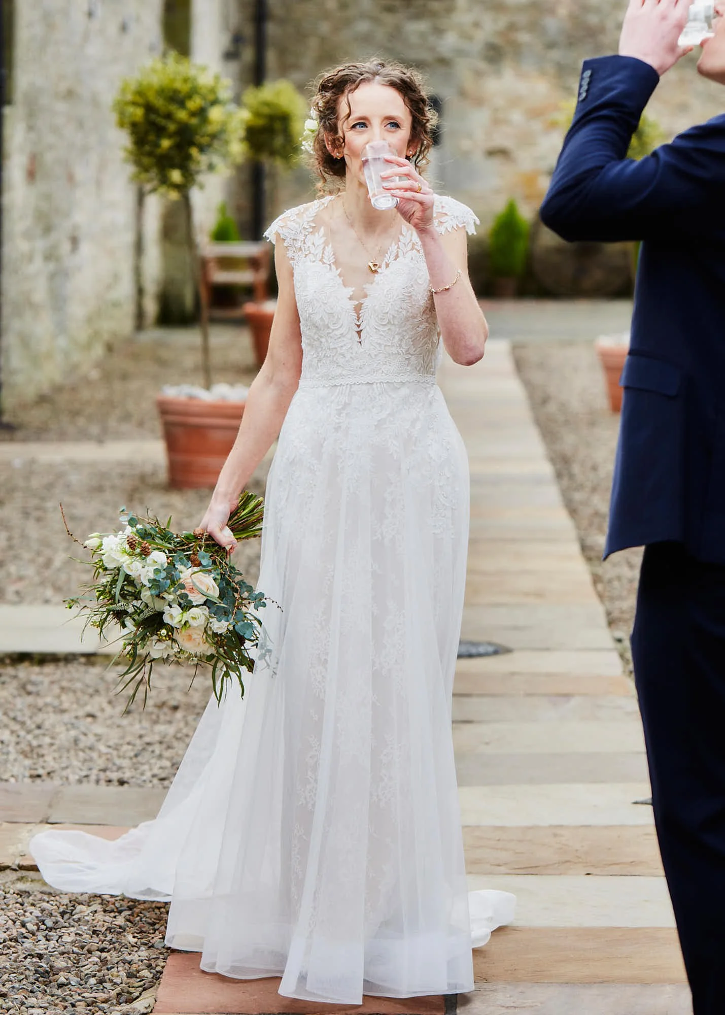 A bride in a white lace wedding dress holding a bouquet of flowers while drinking a glass of water during a wedding ceremony outdoors.
