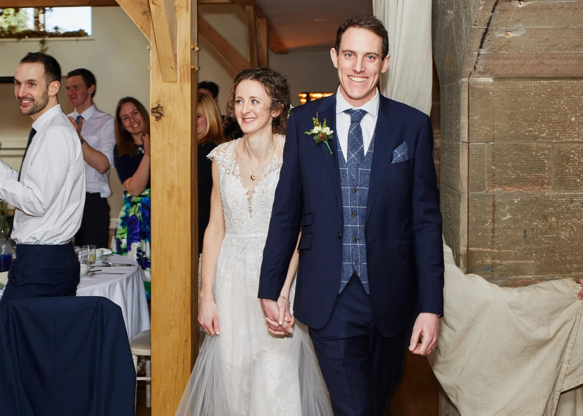 A bride and groom holding hands, standing inside a rustic wedding venue with wooden walls and a window in the background. The groom is wearing a navy blue suit with a plaid vest and a boutonniere, and the bride is wearing a lace white wedding dress w