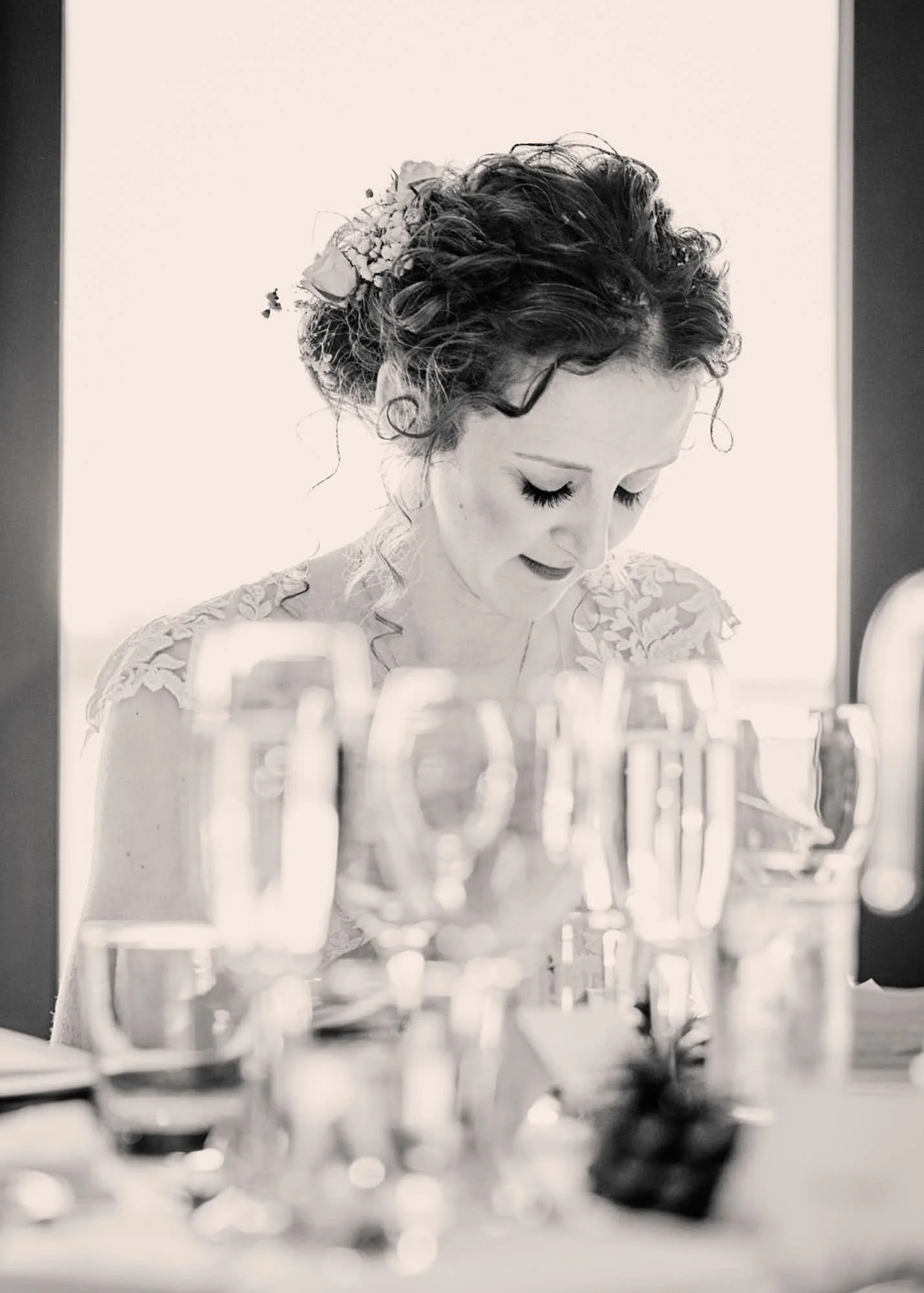 Black and white photo of a woman with curly hair and floral hair accessory, looking down, with blurred glasses and bottles in the foreground.