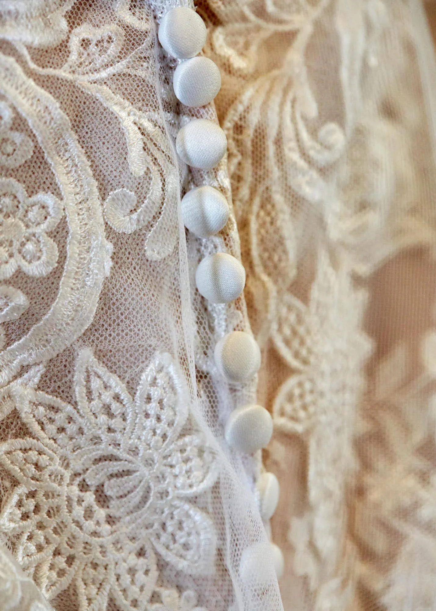 Close-up of cream-colored lace fabric with embroidered floral patterns and a row of fabric-covered buttons.