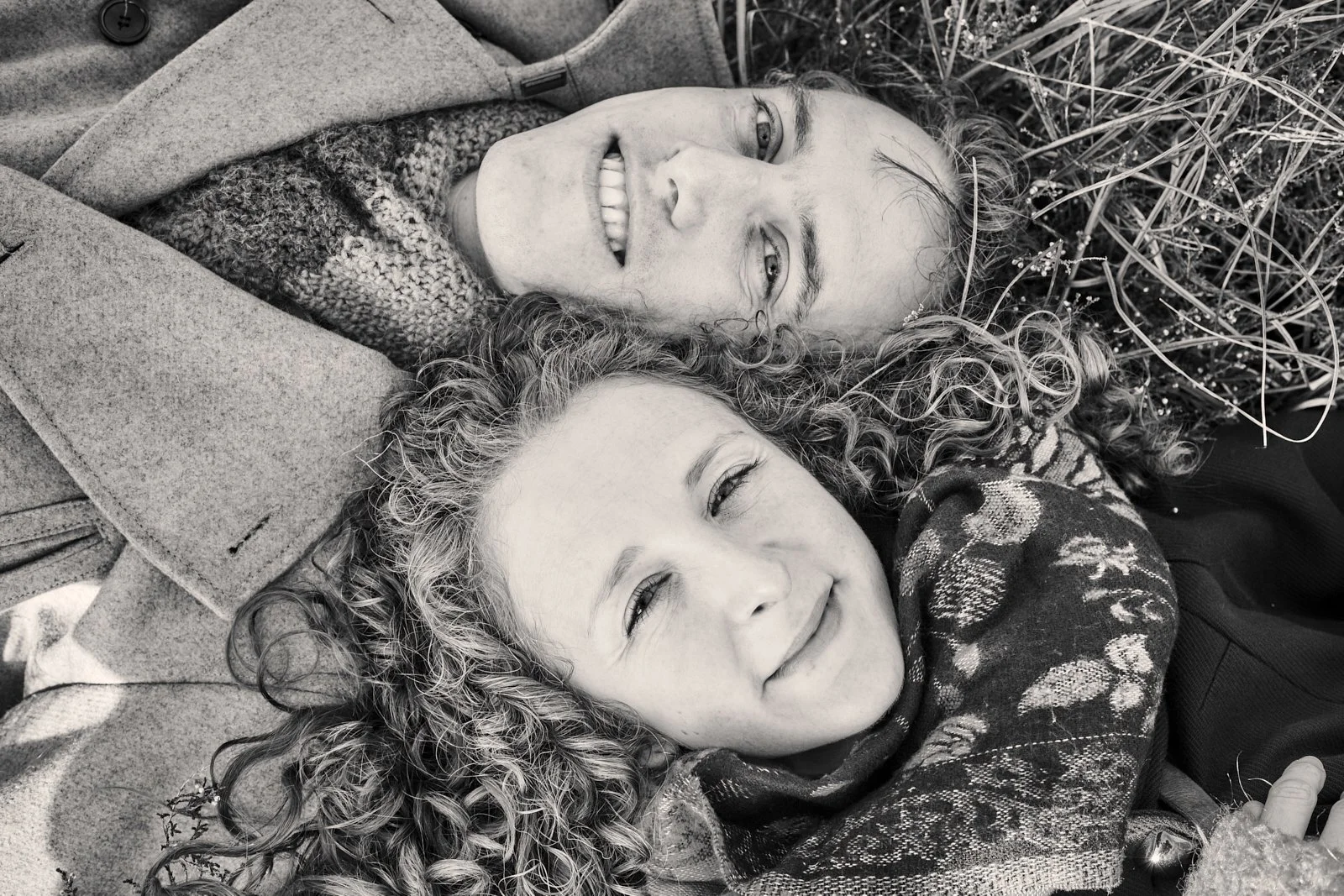 Two women with curly hair lying on the ground among grass and small plants, smiling and looking up.