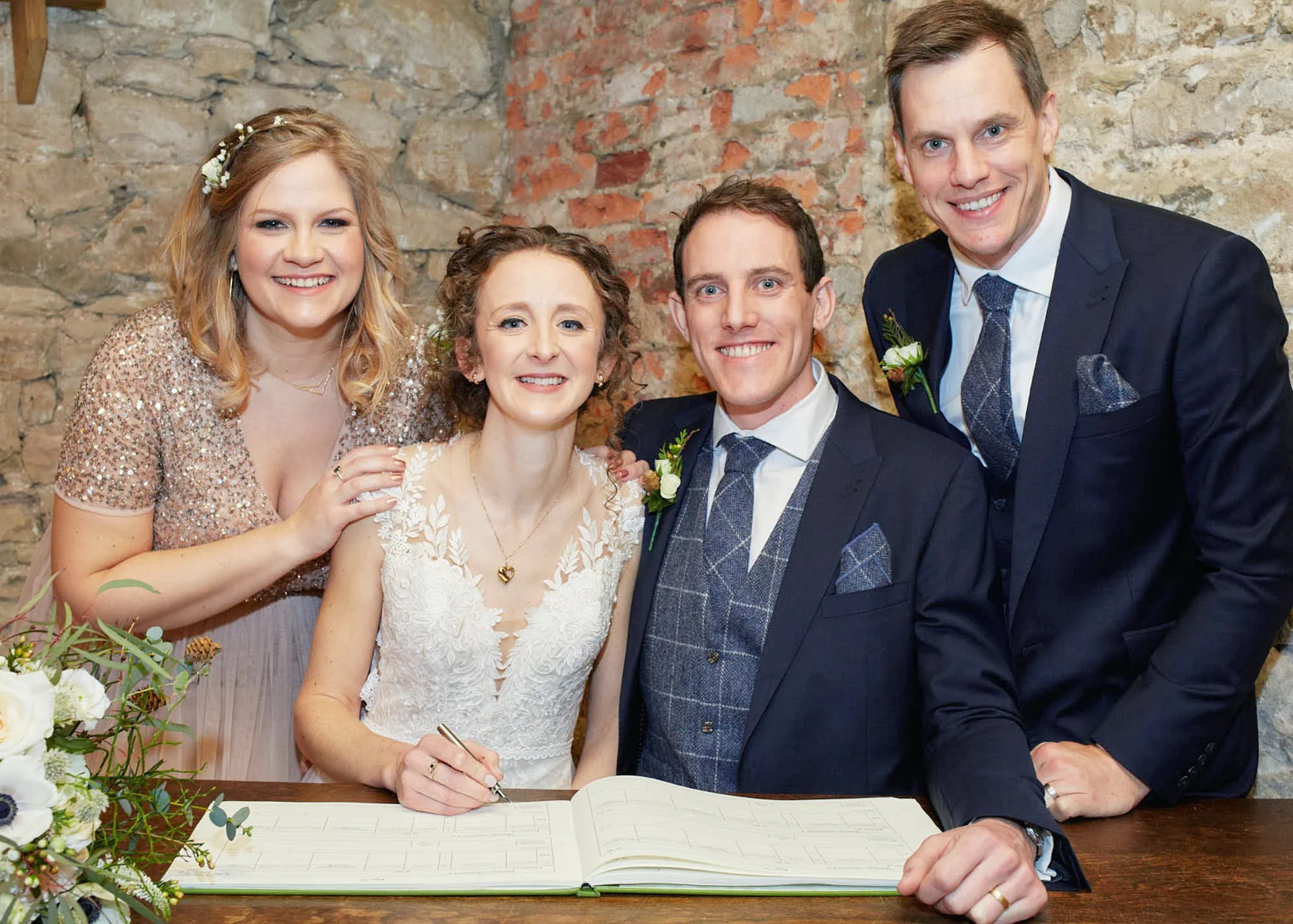 A group of five people at a wedding, with a woman in a white dress signing a guest book, surrounded by smiling friends and family, against a rustic brick wall background.