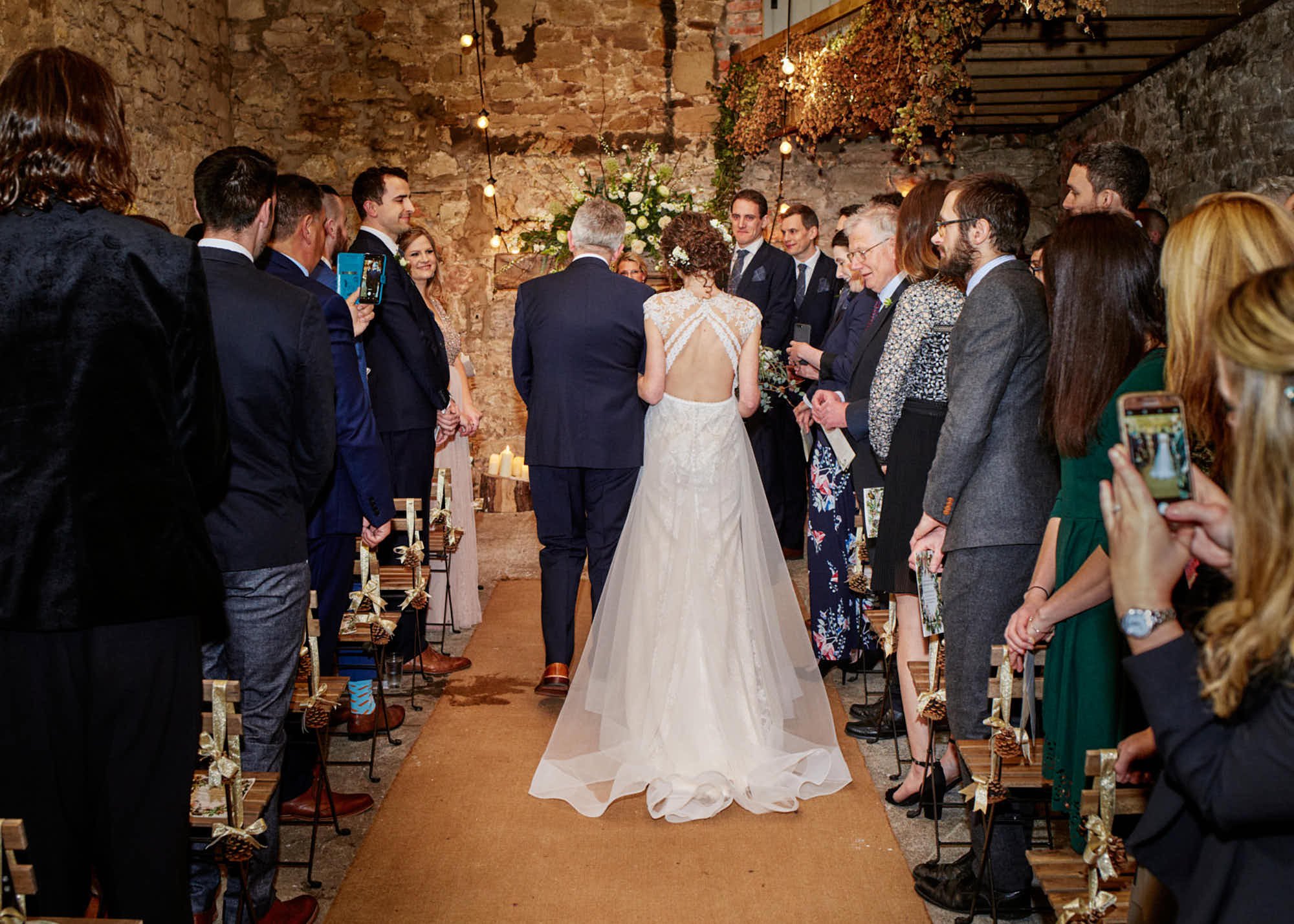 Bride and groom standing at the altar during a wedding ceremony, surrounded by seated guests in a rustic indoor venue with stone walls and floral decorations.