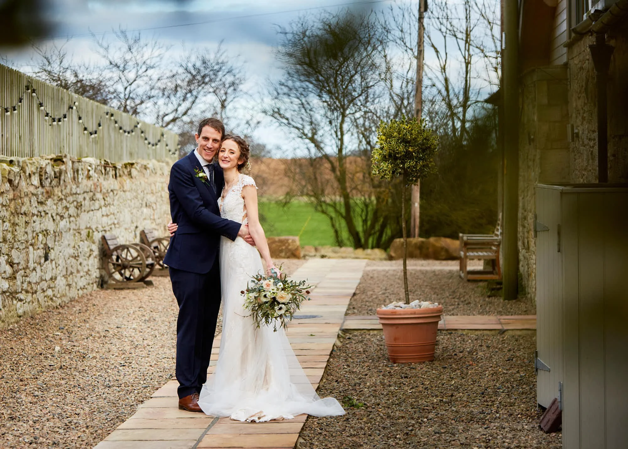 A newlywed couple stands outdoors in front of a stone patio and a wooden fence with string lights, surrounded by leafless trees and a cloudy sky, smiling and embracing.