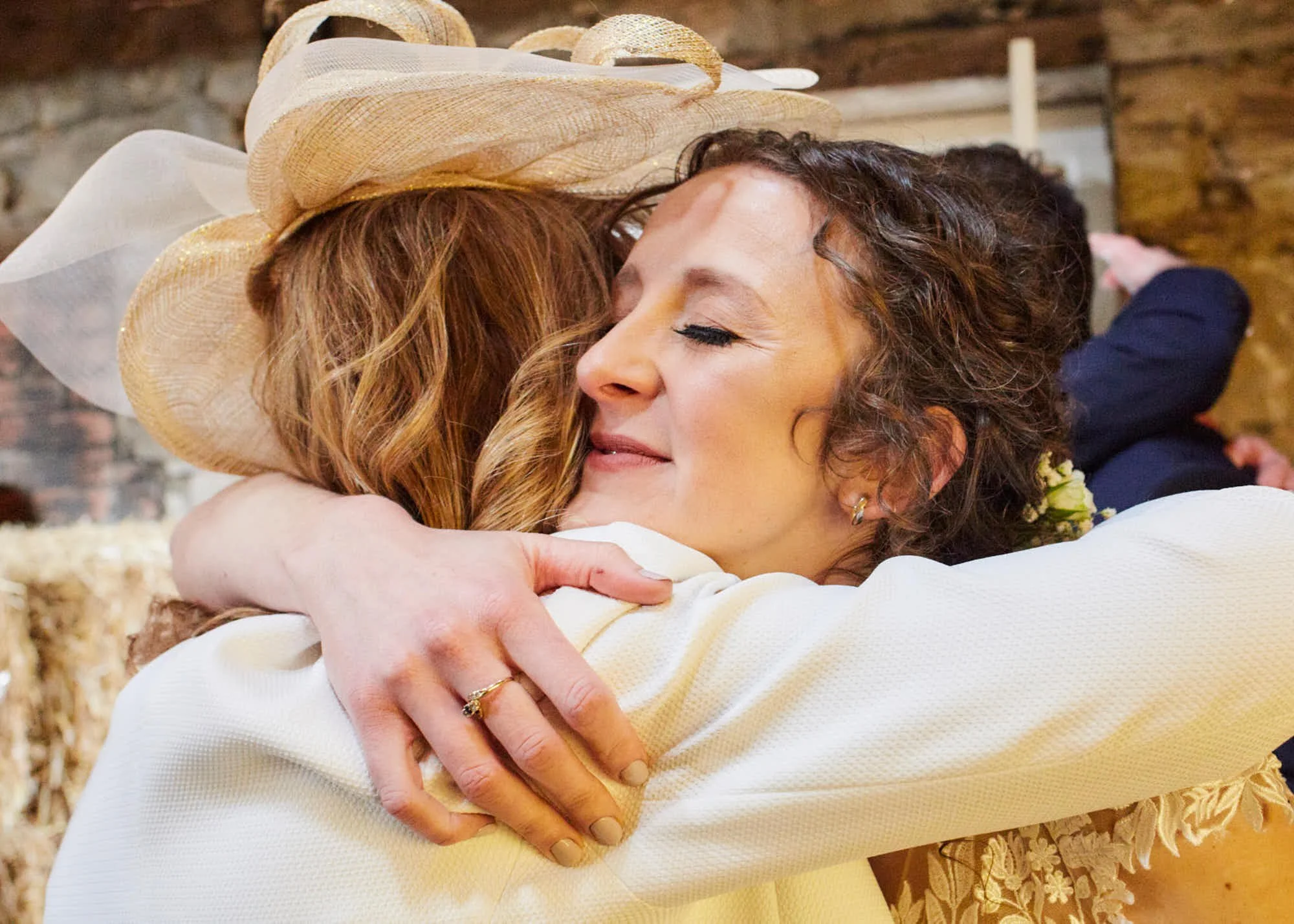 Two women hugging warmly, one wearing a fancy hat, inside a rustic setting.