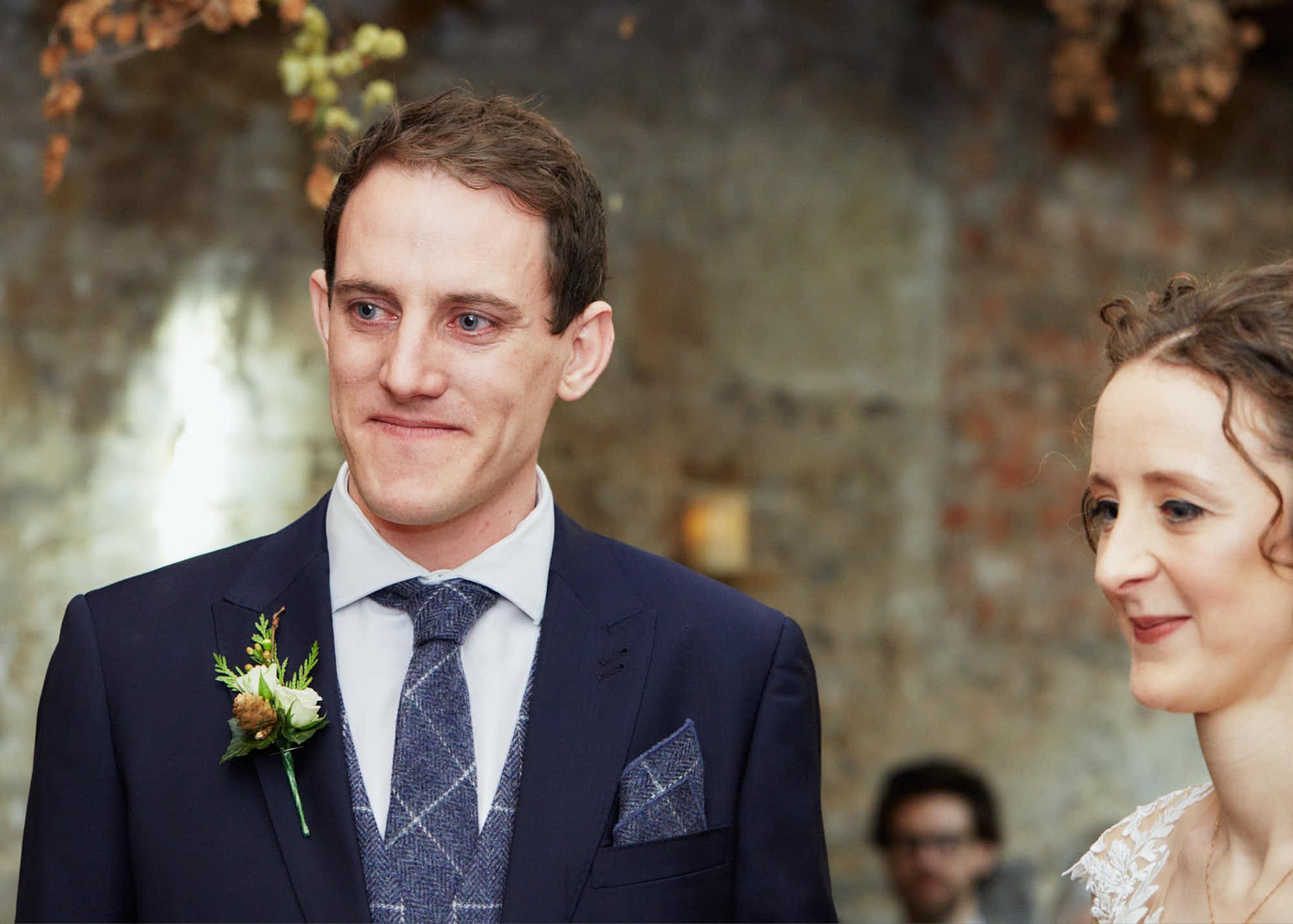 A groom in a navy suit, white shirt, and patterned tie, standing outdoors with a woman, possibly during a wedding ceremony. The groom has a boutonnière on his left lapel and has short brown hair and light blue eyes. The woman is wearing lace and has 