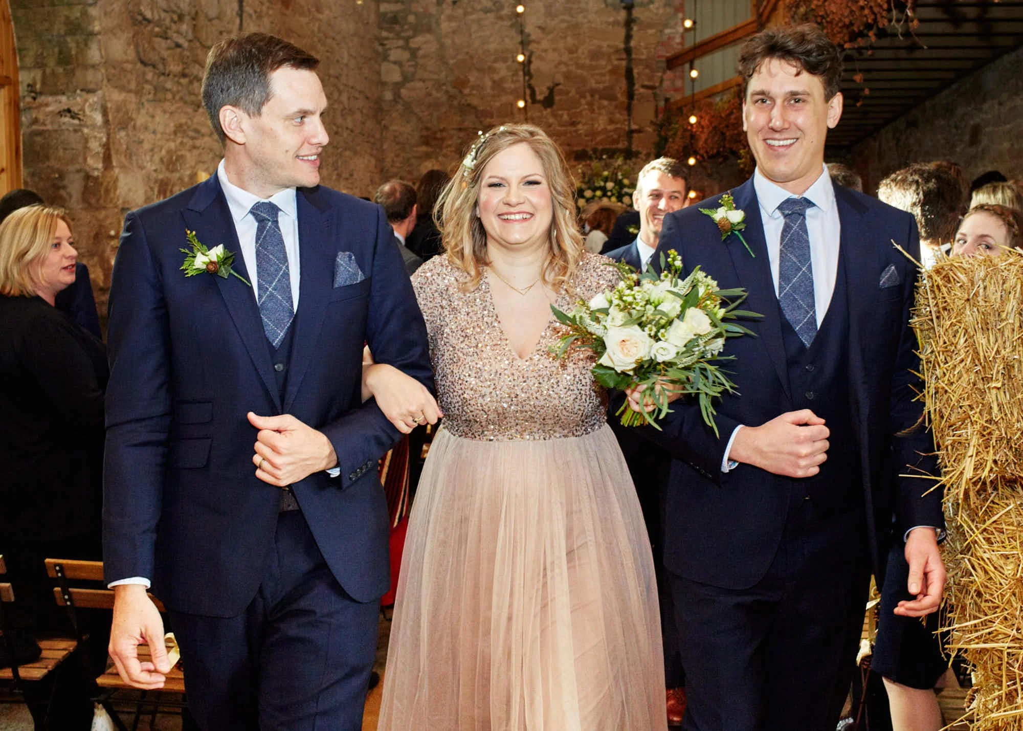 A woman holding a bouquet of white roses, standing between two men in suits, at a wedding reception.