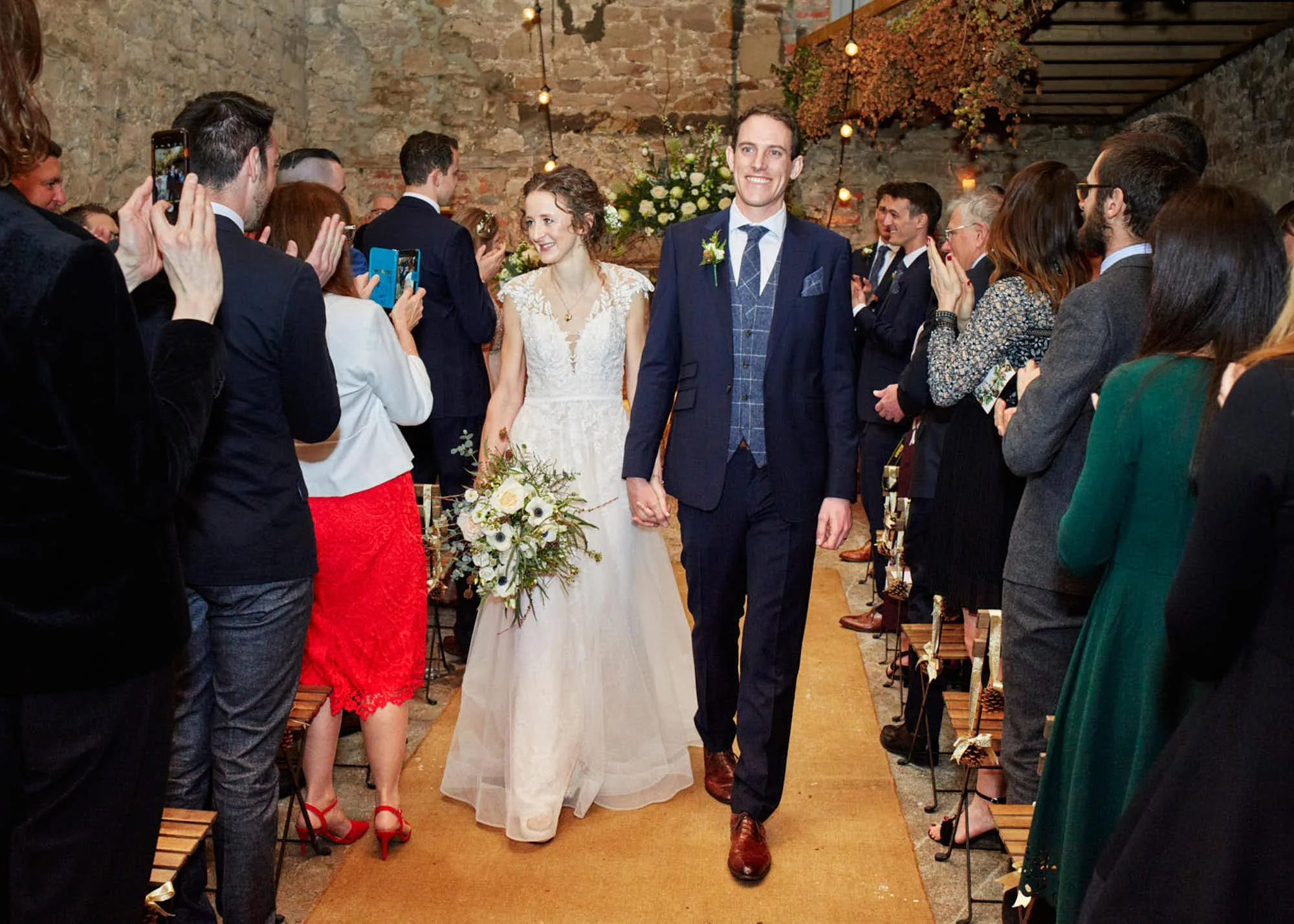 A couple, a bride and groom, walking hand in hand down an aisle during a wedding ceremony surrounded by friends and family in a rustic indoor venue.