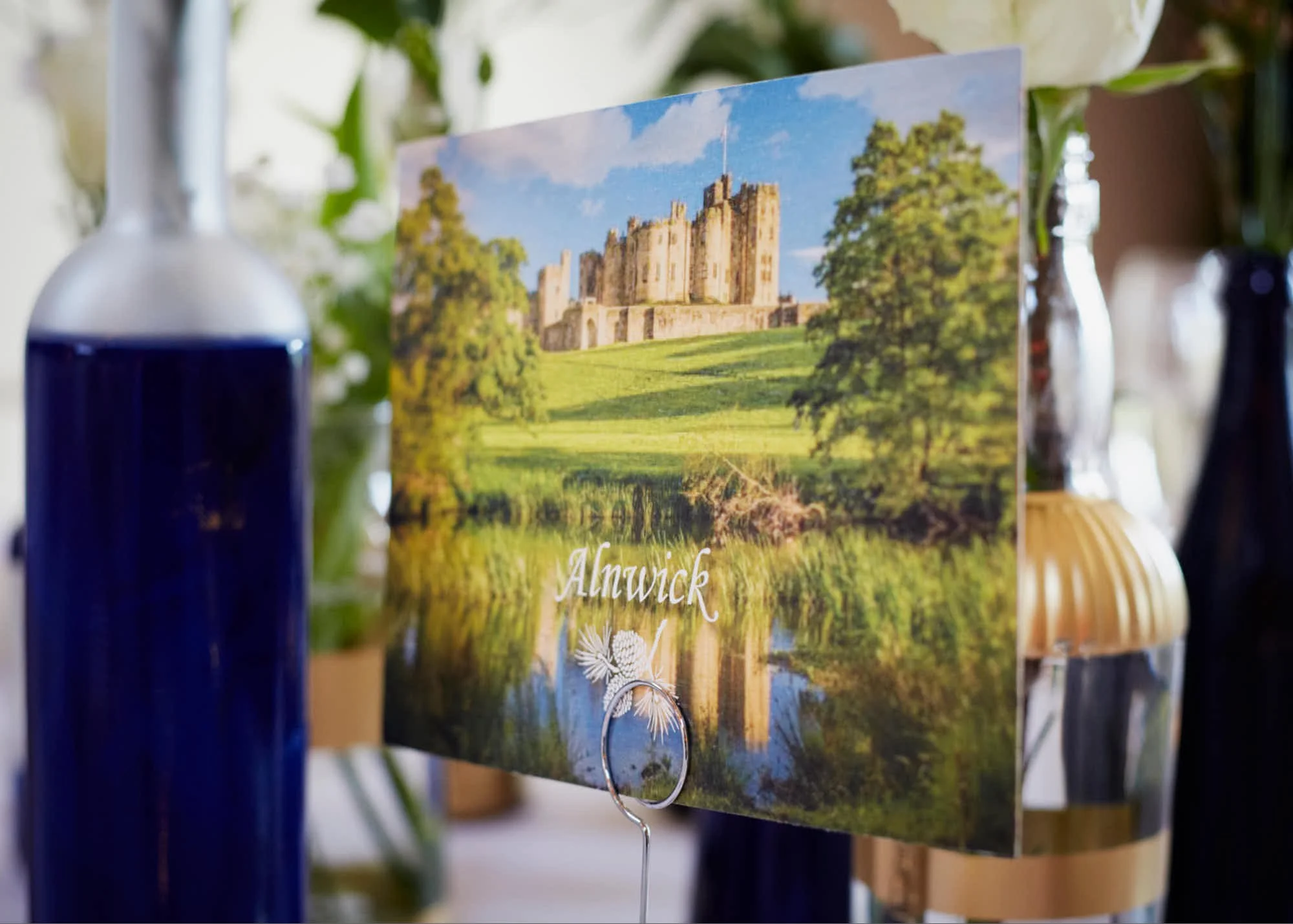 A postcard featuring a castle with a green lawn and a blue sky, surrounded by trees, with the text 'Anwick' on it.