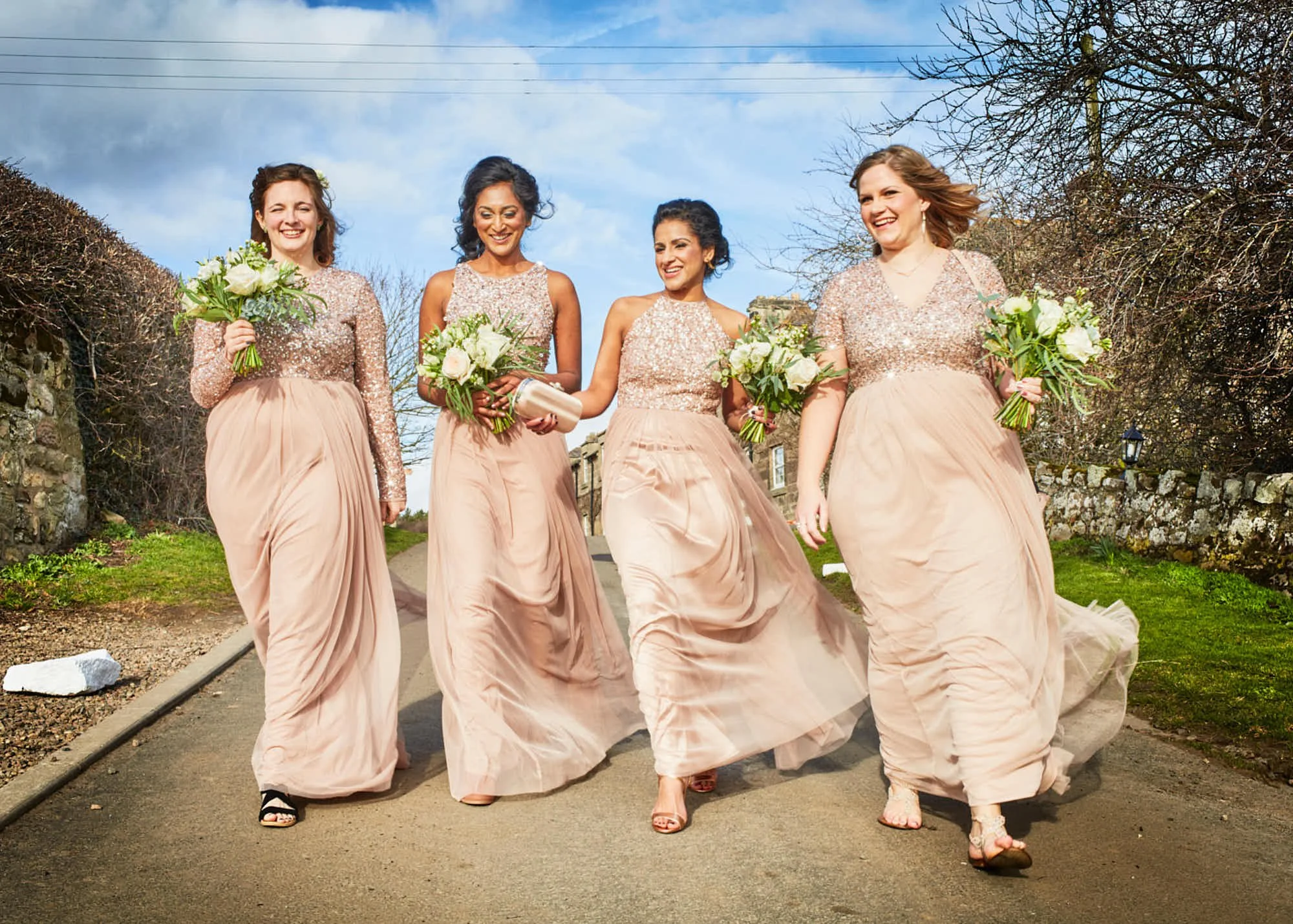 Five women in matching pink strapless dresses walking along a street, holding bouquets, smiling, with trees and a stone wall in the background.