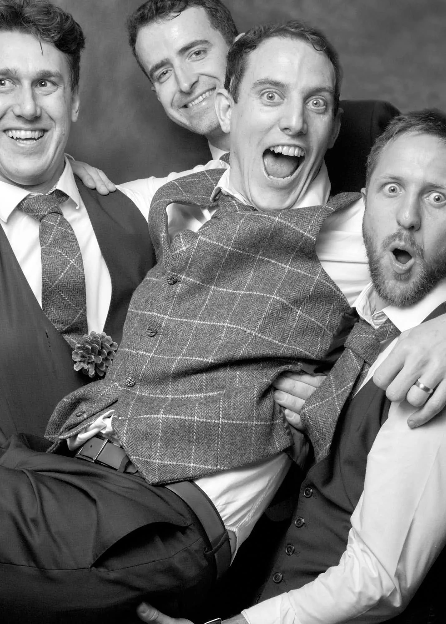 Black and white photo of five smiling men in formal attire posing together, with one man in the center showing a surprised expression, others smiling widely, all closely gathered and appearing cheerful.