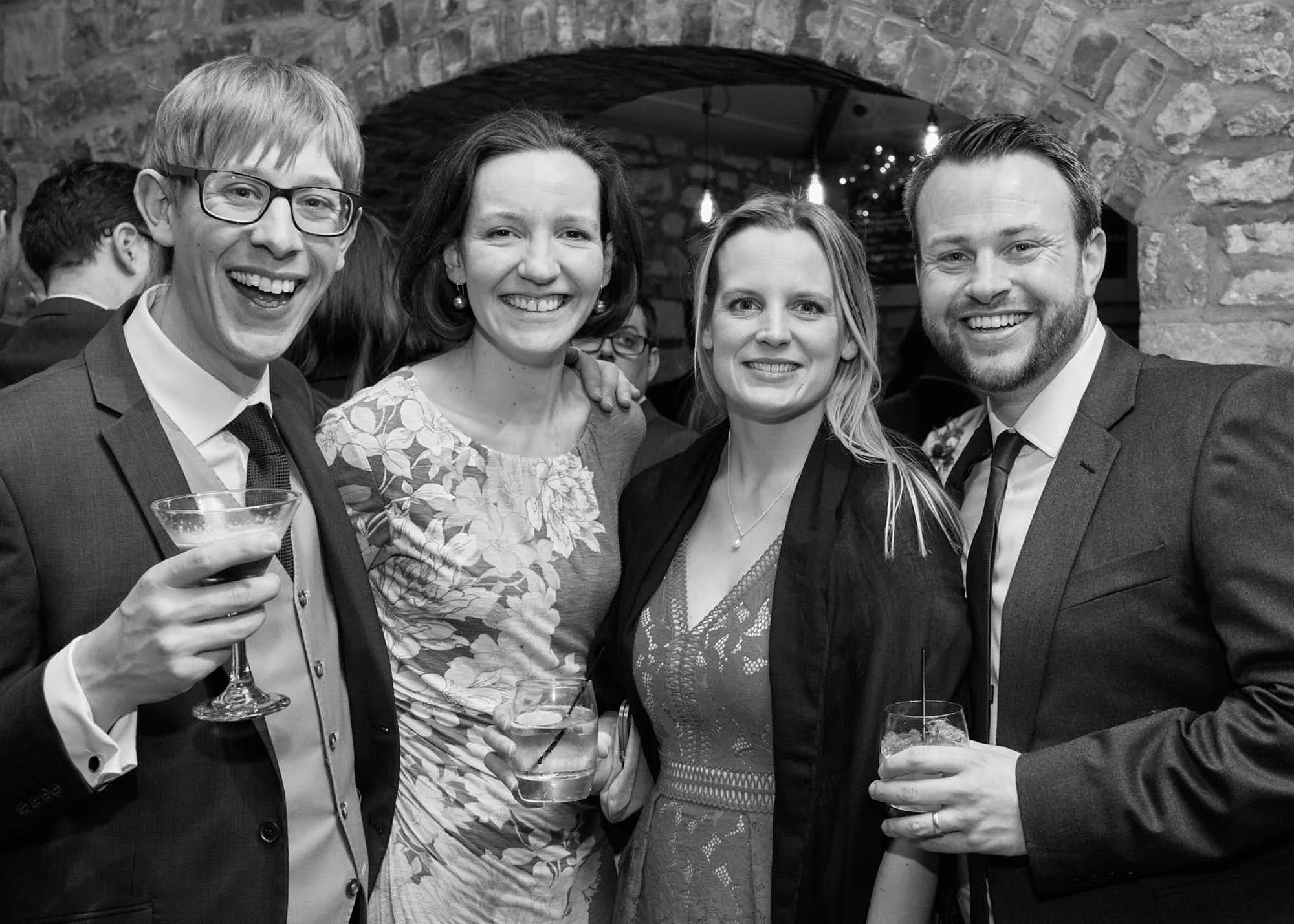 Four people, two men and two women, smiling and holding drinks at a party or celebration in a brick-walled venue.