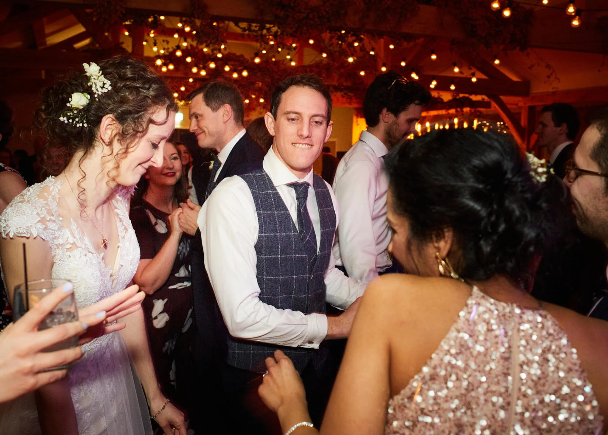 People dancing and socializing at a wedding reception or party with warm lighting and hanging string lights.