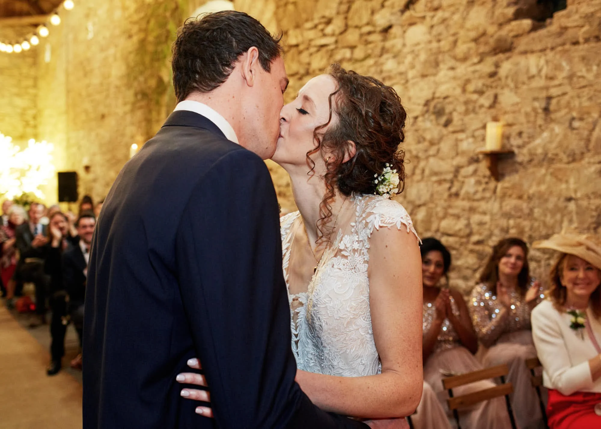 A bride and groom share a kiss during their wedding ceremony, with wedding guests seated and watching in the background at an indoor venue with a brick wall.
