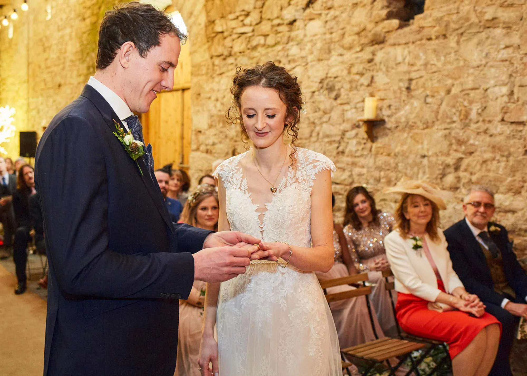 A bride and groom exchanging rings during their wedding ceremony in a rustic venue with exposed brick walls. Guests are seated and watching the couple.