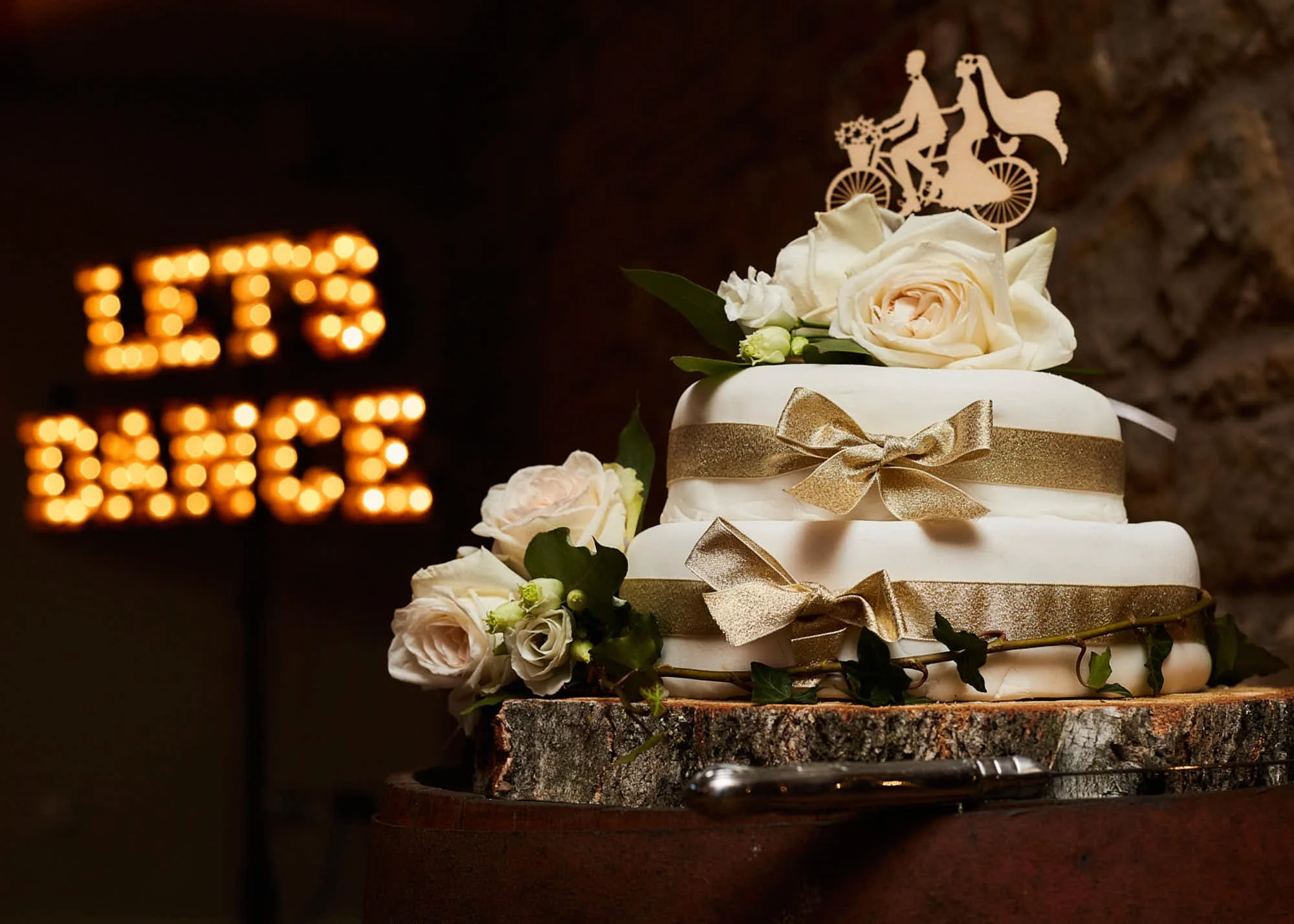 A wedding cake decorated with white roses, greenery, and gold ribbons, with a cake topper of a bride and groom riding a tandem bicycle, set on a rustic wooden surface.