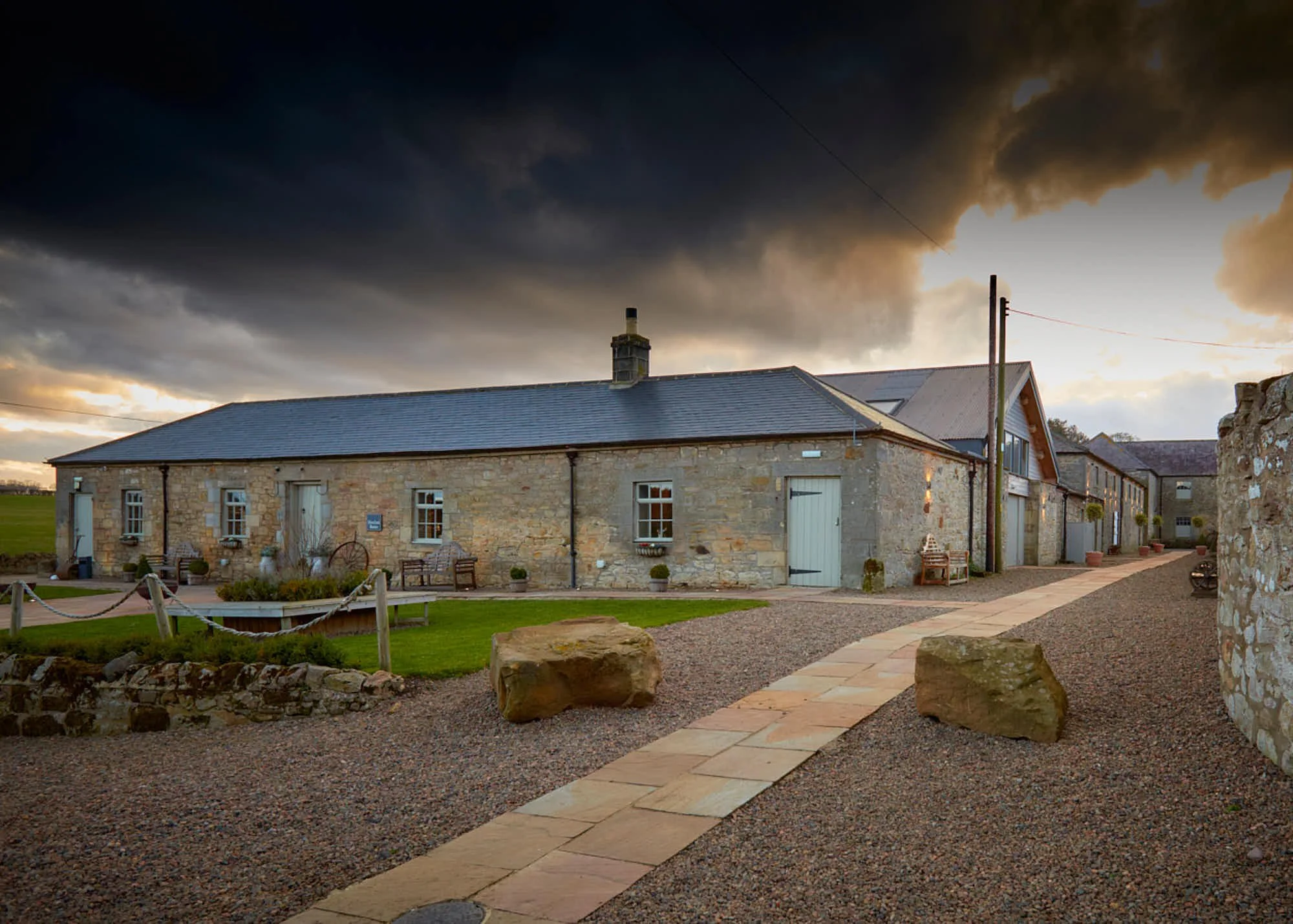 Stone building with a metal roof and a chimney, cloudy sky, and a stone-paved walkway in the foreground.