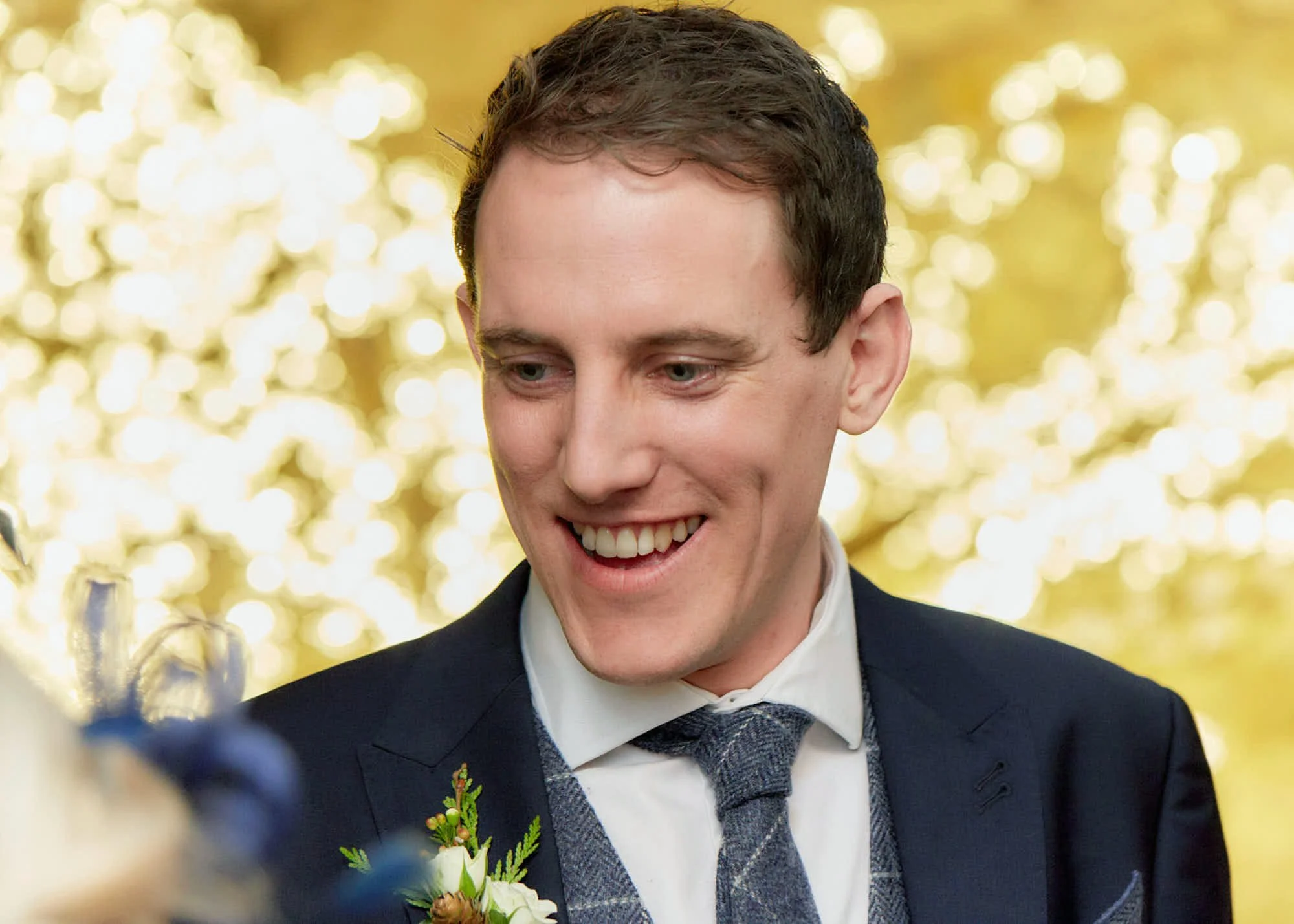 A young man with short brown hair, dressed in a dark suit with a boutonniere, smiling with his teeth visible, at a celebration or formal event with blurred golden lights in the background.