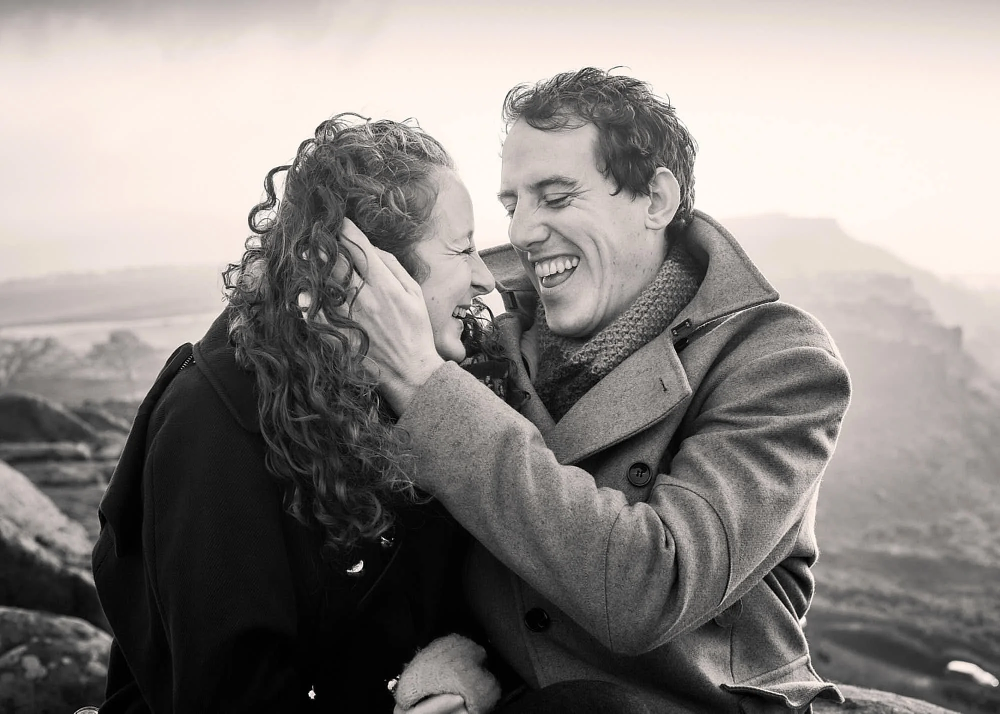 A black-and-white photograph of a couple outdoors, smiling and embracing each other with their foreheads touching, in a nature setting with rocks and mountains in the background.