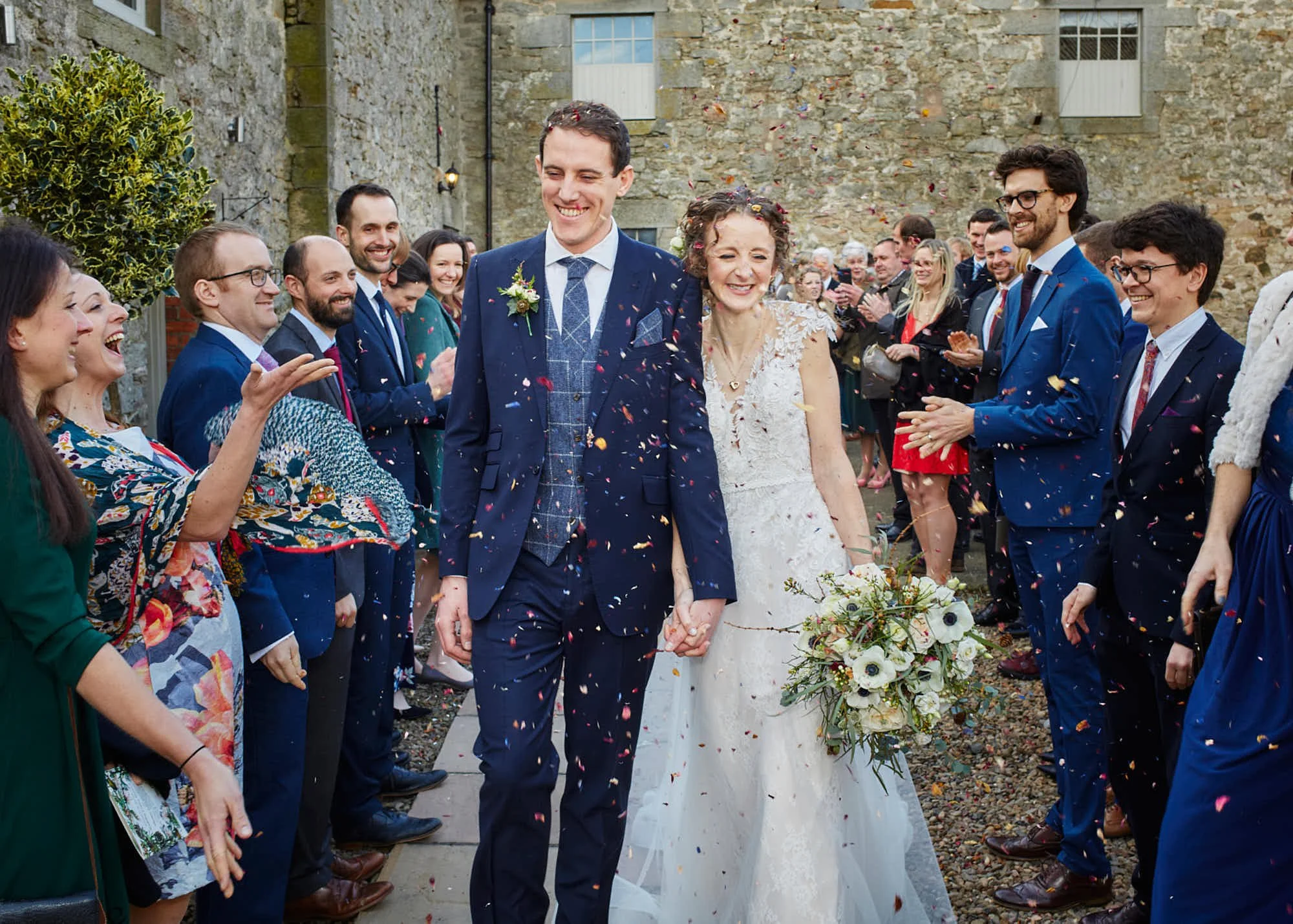 A wedding scene outside a stone building shows a smiling bride and groom holding hands, surrounded by friends and family clapping, cheering, and throwing confetti.