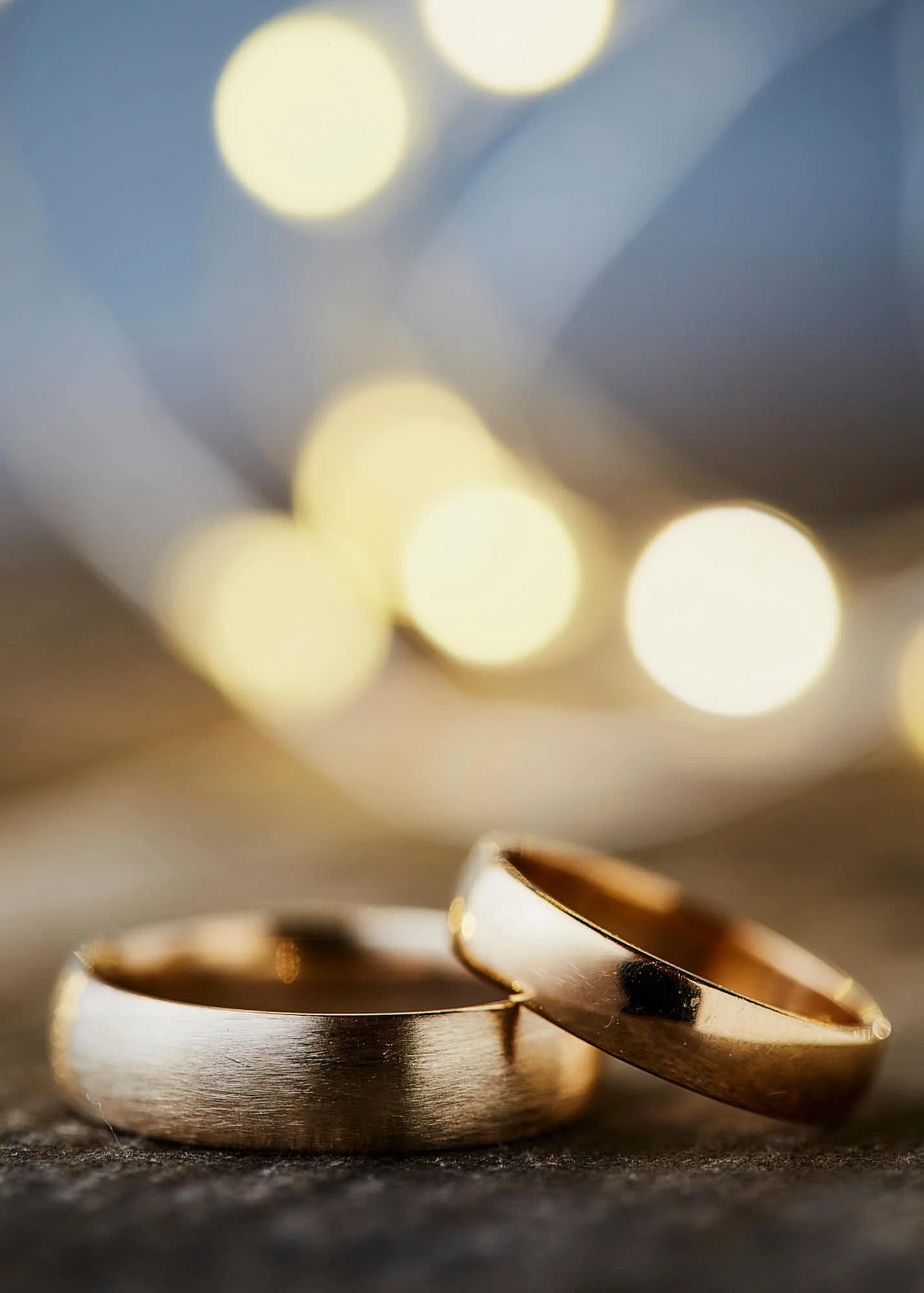 Close-up of intertwined gold wedding rings resting on a surface, with a blurred background of warm lights.