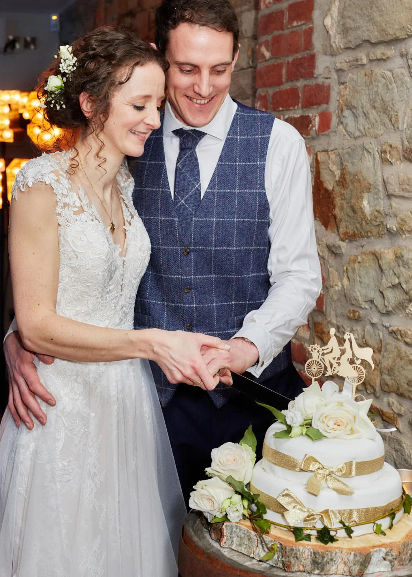 A bride and groom cutting a wedding cake decorated with white roses and gold ribbons at their wedding reception.