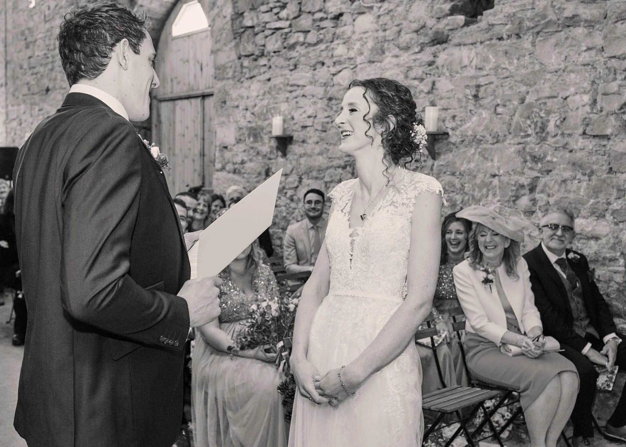 A bride and groom exchanging vows during a wedding ceremony inside a rustic stone building with guests seated and smiling in the background.