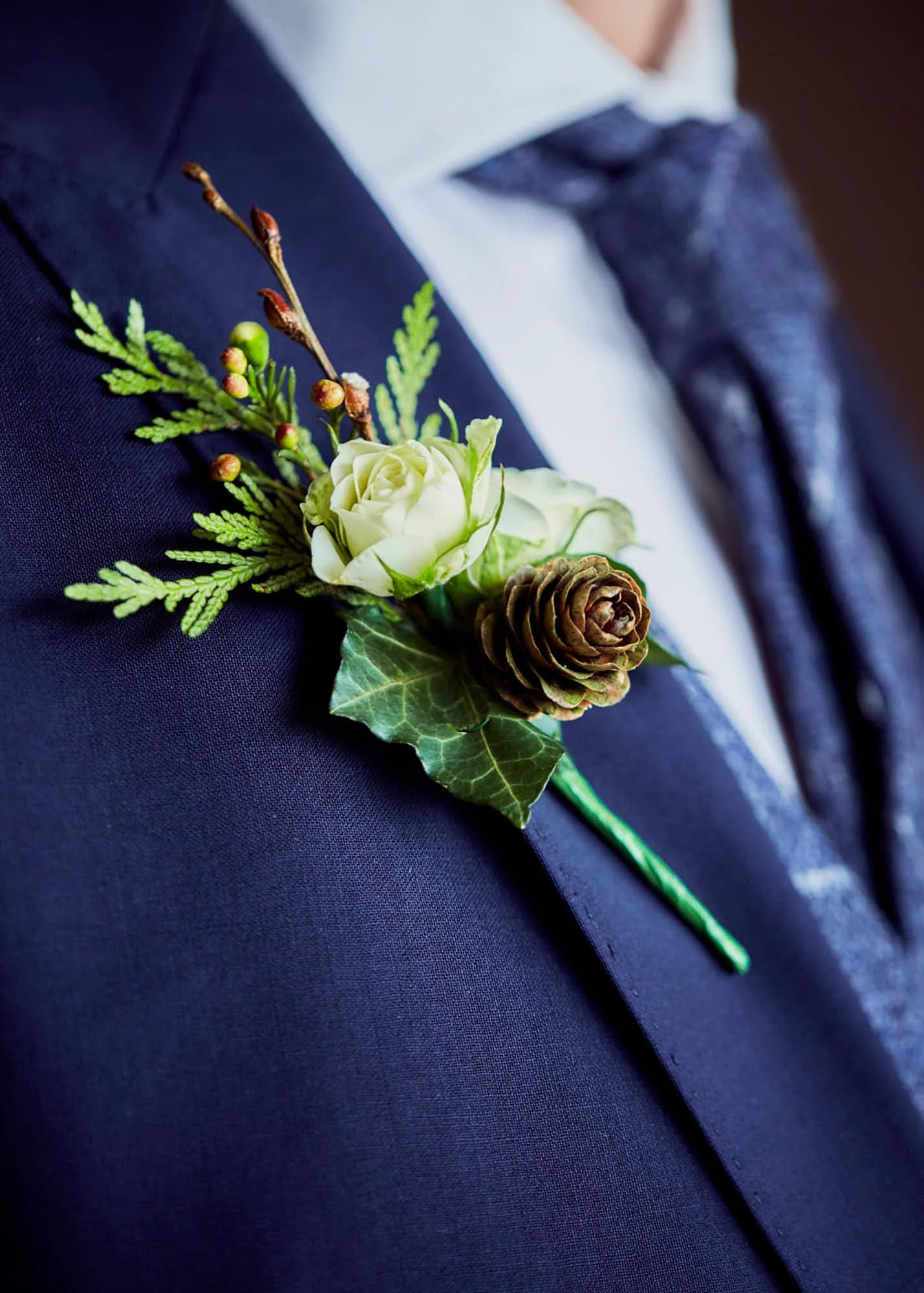 Close-up of a boutonniere pinned to a navy blue suit jacket, featuring a white rose, pine needles, a pinecone, and holly leaves.