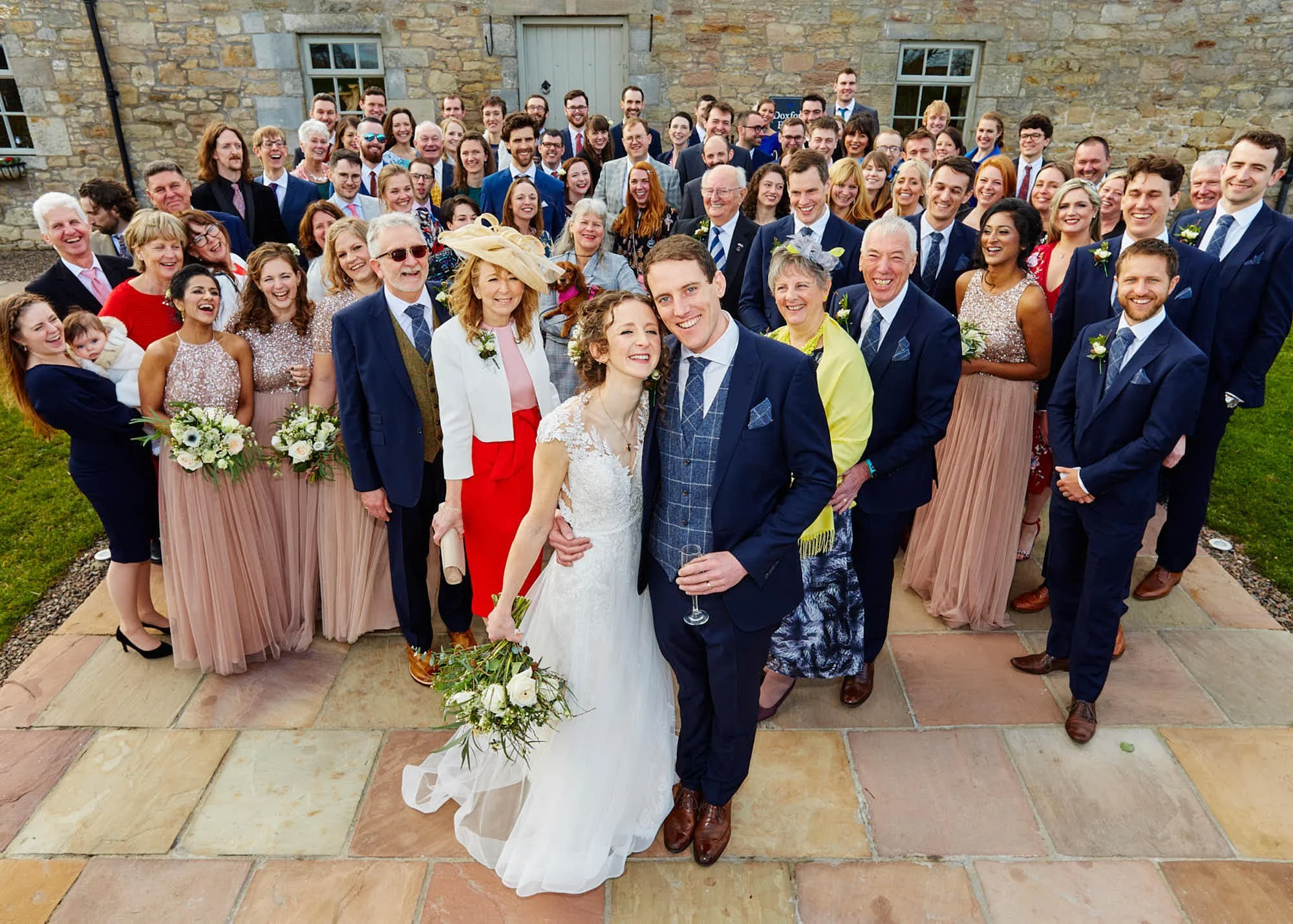 A large group of people, dressed in formal attire, gathered outdoors in front of a stone building for a wedding celebration. The bride and groom are in the front center, smiling and holding a bouquet of flowers, with the groom also holding a glass of