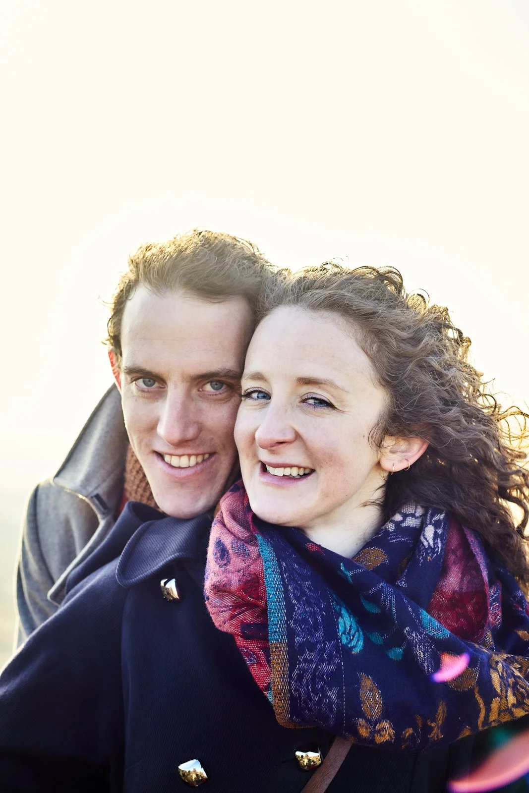 Close-up of a smiling young man and woman embracing outdoors, bright sunlight background.
