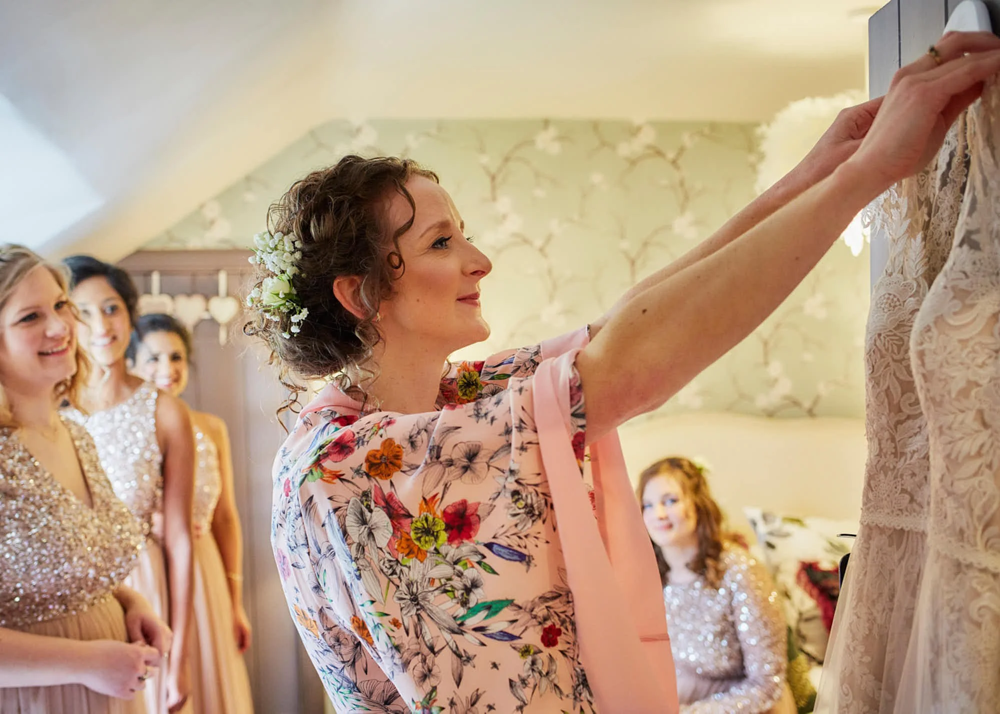 Woman in floral pink robe adjusting lace dress on mannequin at a wedding or event. Other women in formal dresses watching smiling.