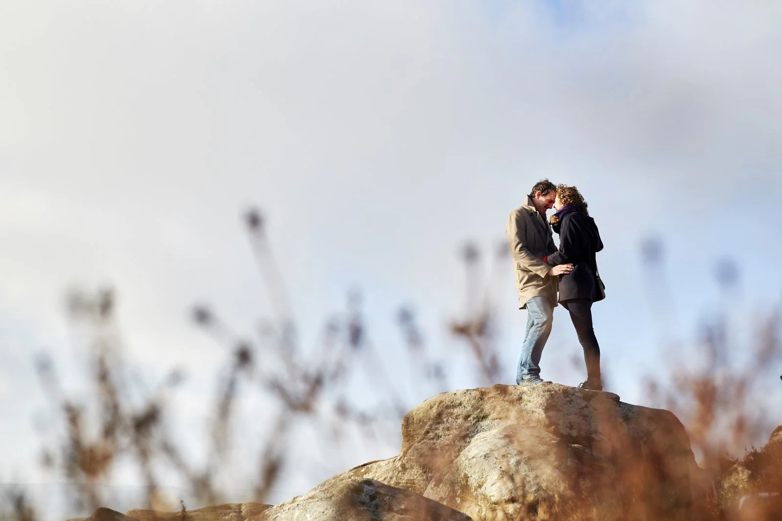 A couple standing close together on a rock, sharing a moment and holding hands, with a cloudy sky background and blurred foreground plants.