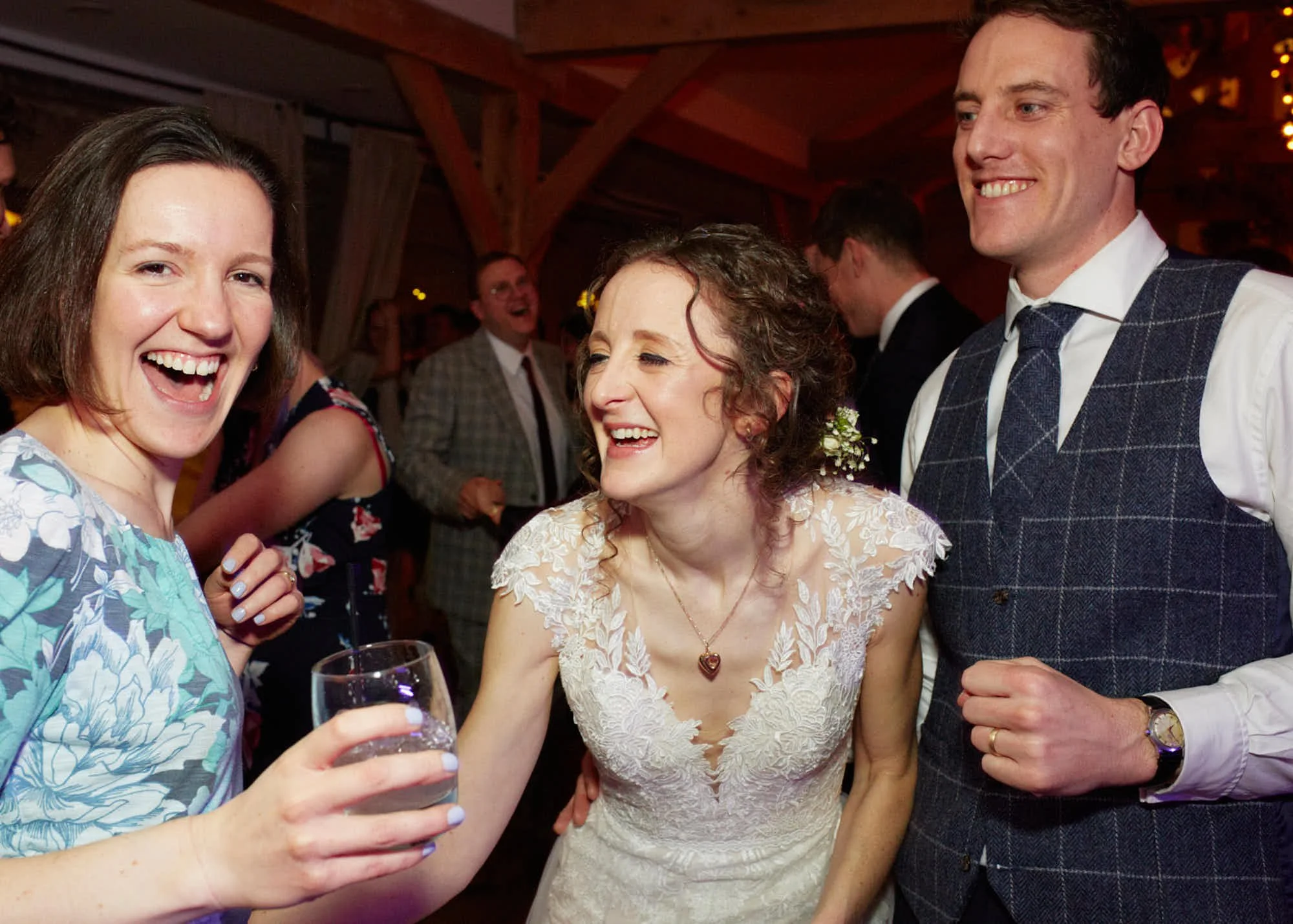 Group of people enjoying a celebration indoors, smiling and holding drinks. Woman in white lace dress at the center, surrounded by smiling friends.
