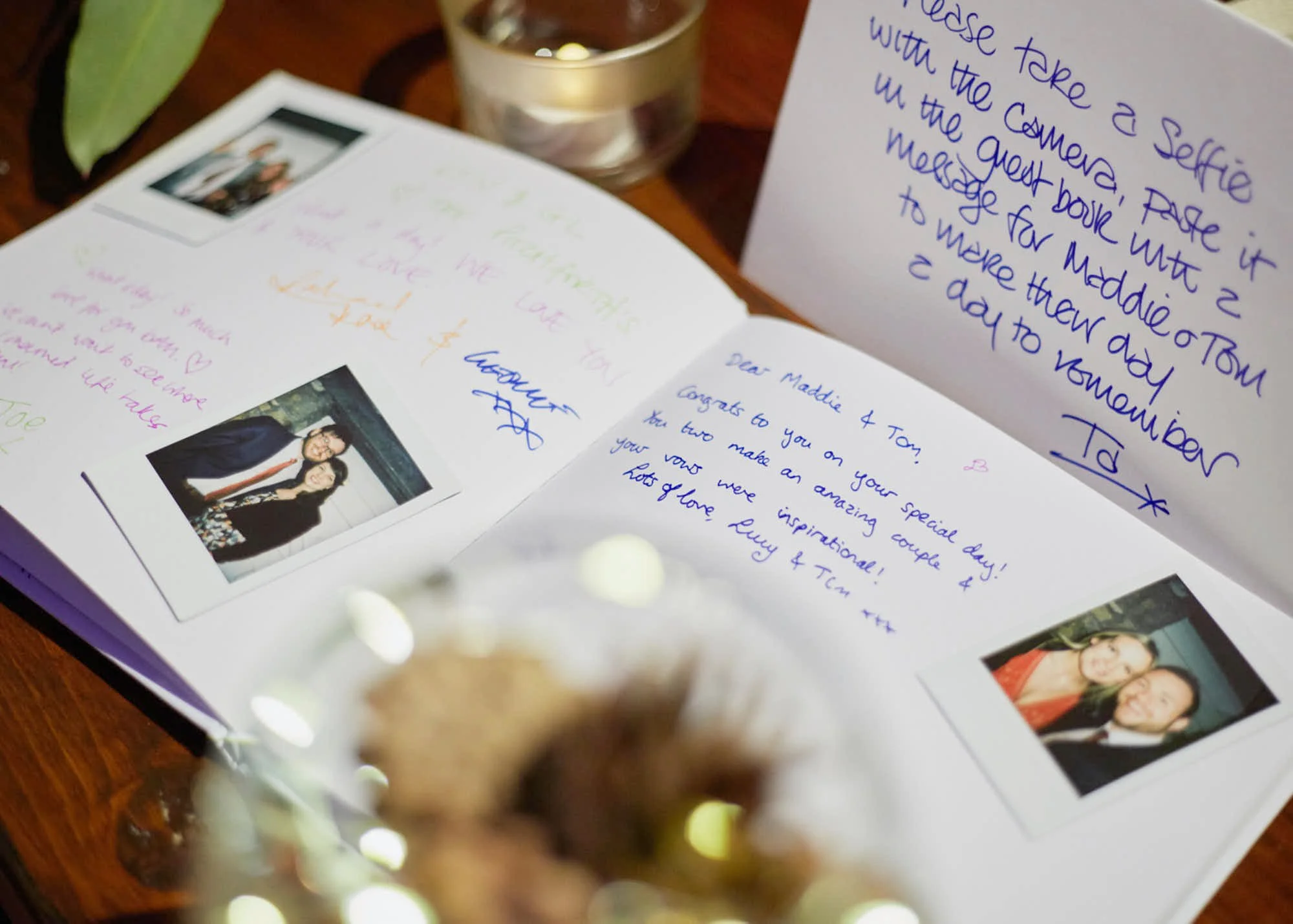 Open greeting card with handwritten messages and photo of two women, on a wooden surface with a candle and blurred decorative objects in the background.