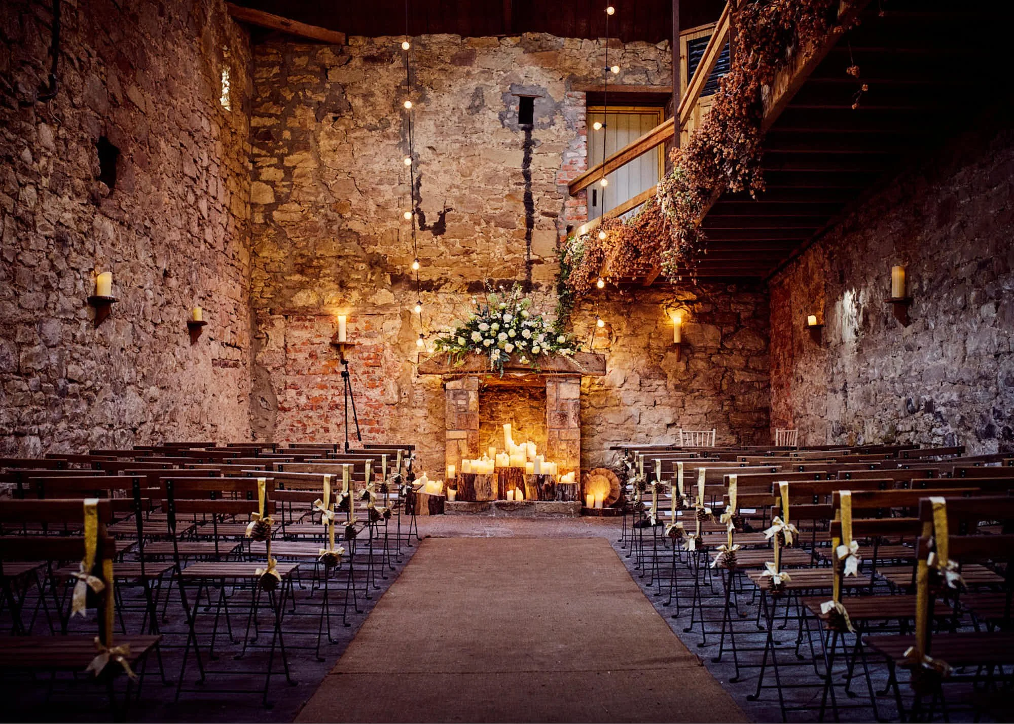 Indoor wedding ceremony setup with rows of chairs, a central aisle, candle decorations, and a floral display in a rustic stone-walled venue.