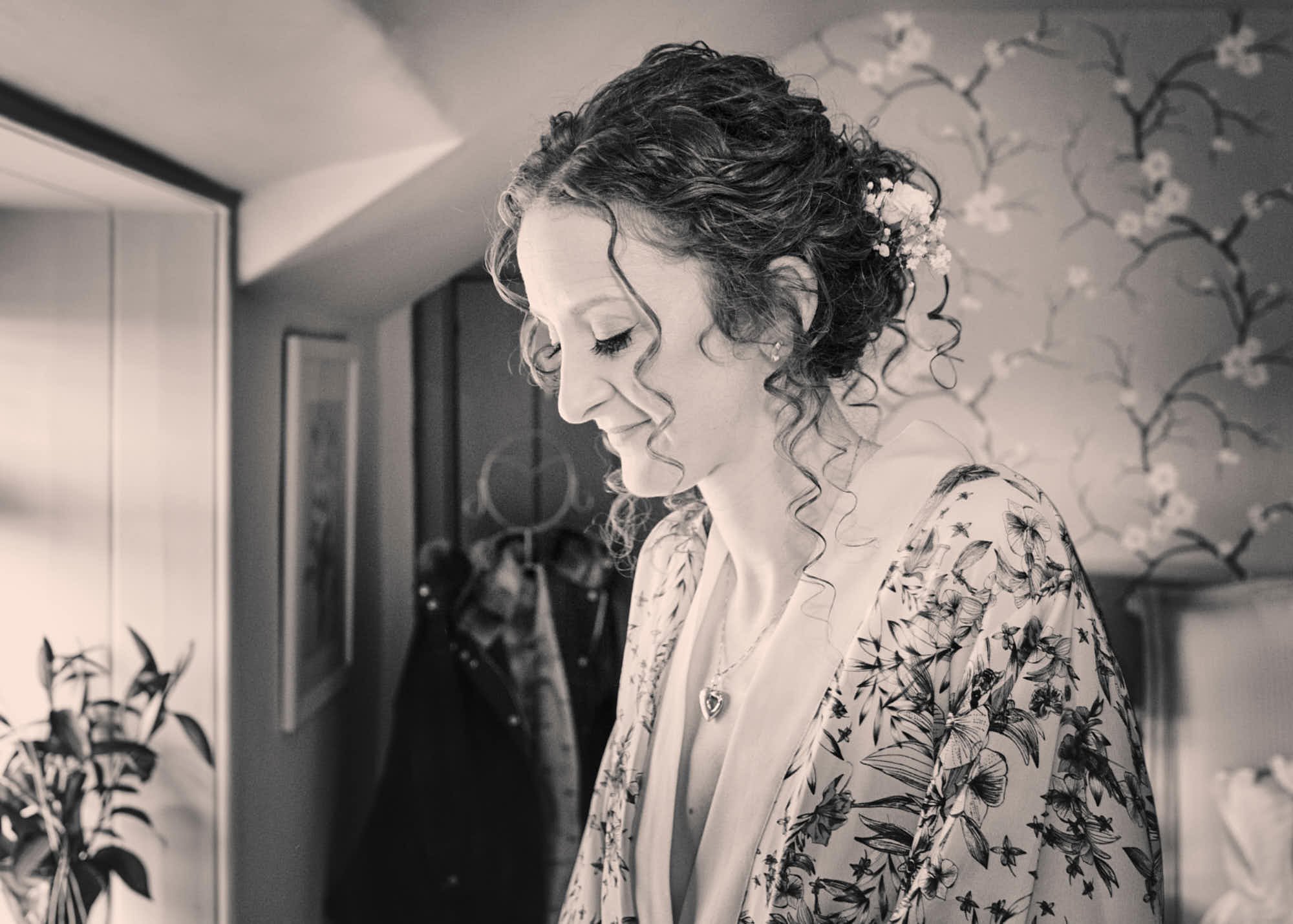 Black and white photo of a woman with curly hair decorated with flowers, looking down with a gentle smile, indoors with floral wallpaper and a mirror.