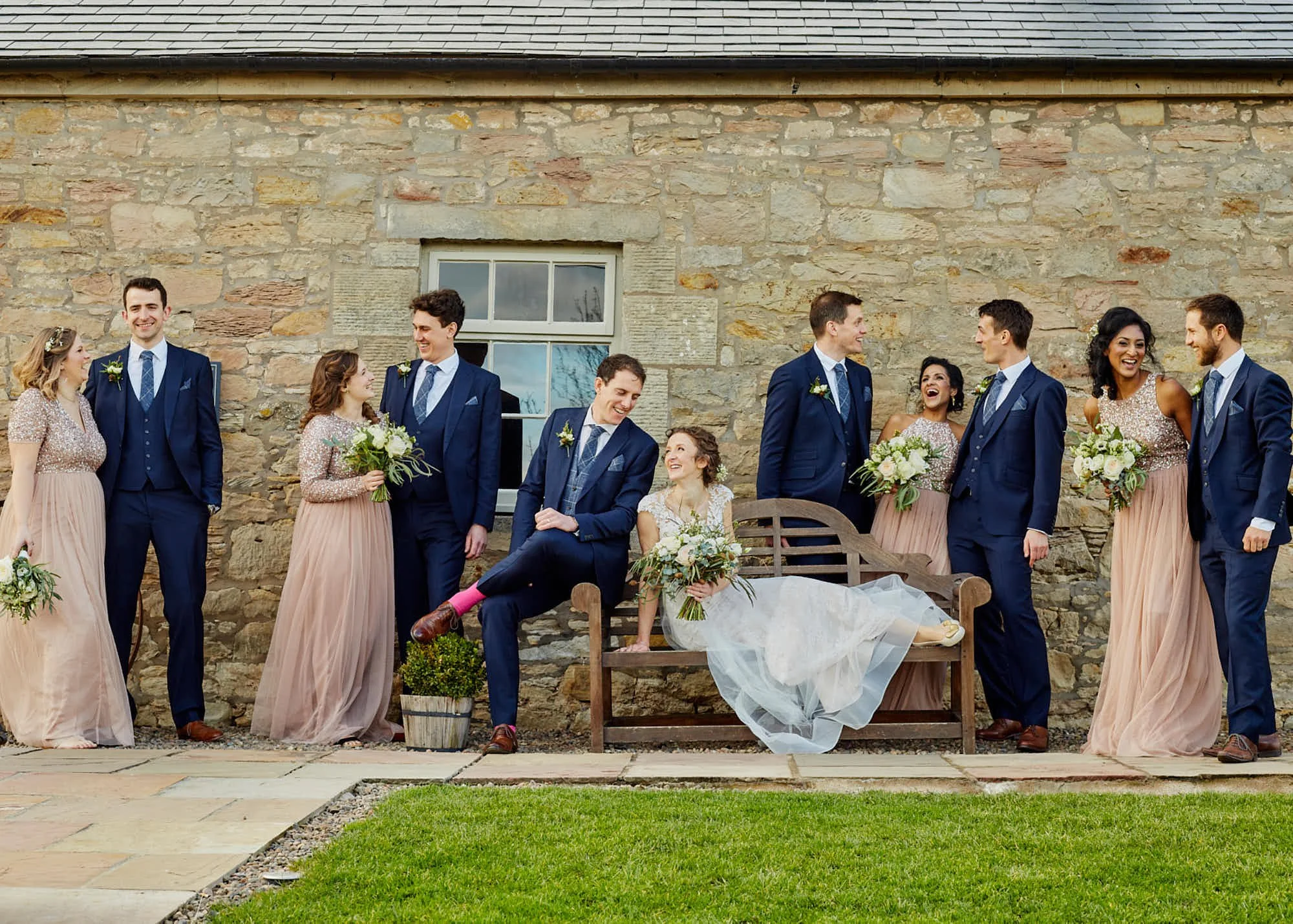 Group wedding party outside in front of a stone building, with bridesmaids in pink dresses holding bouquets, groomsmen with blue suits, and a bride and groom sitting on a bench, all smiling and talking.