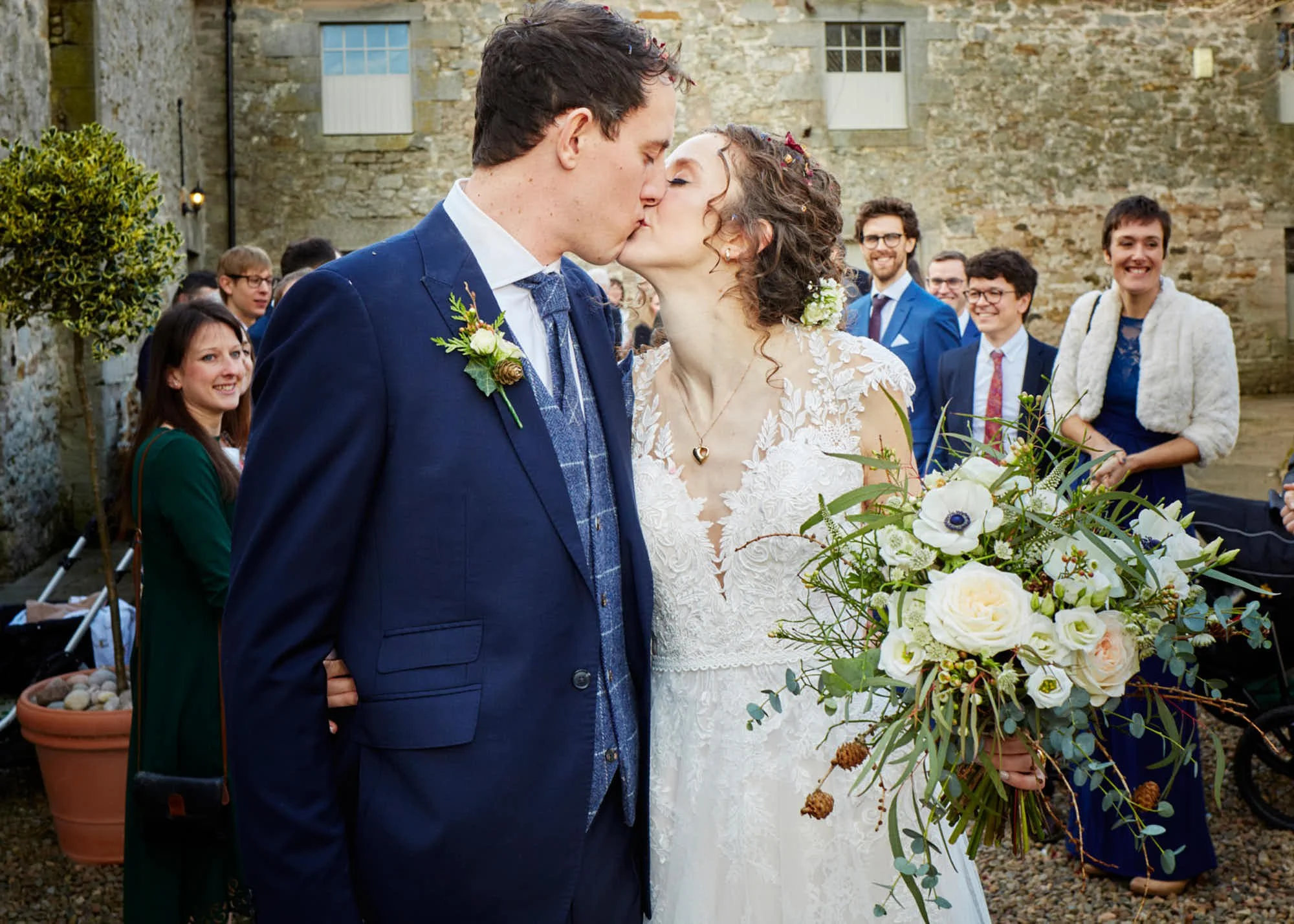 A bride and groom kissing at their wedding ceremony outdoors, with wedding guests in the background smiling, holding flowers, and dressed in formal attire, against a rustic stone wall.