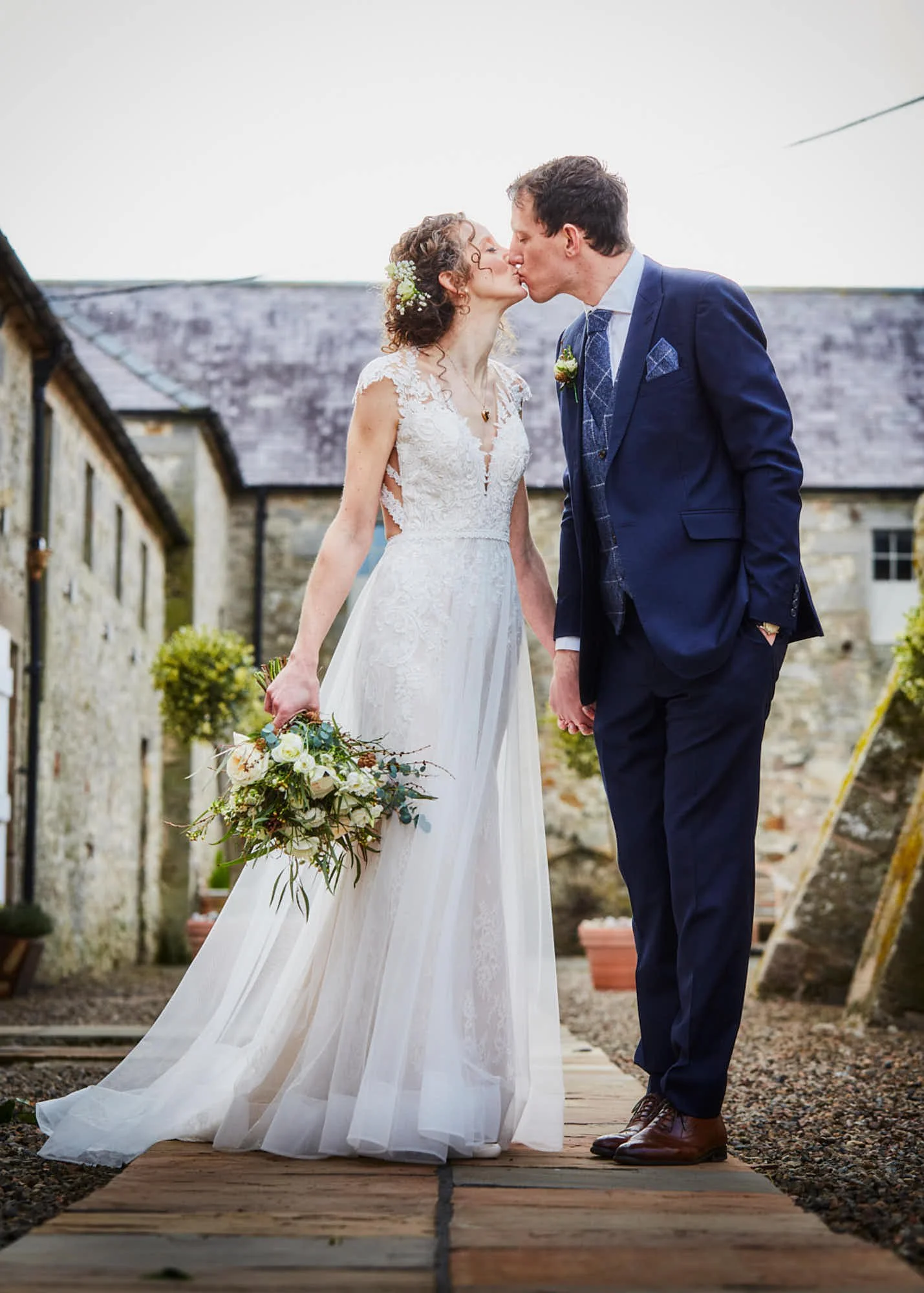 Bride and groom kissing during wedding ceremony outside on a wooden pathway with rustic stone buildings in the background.