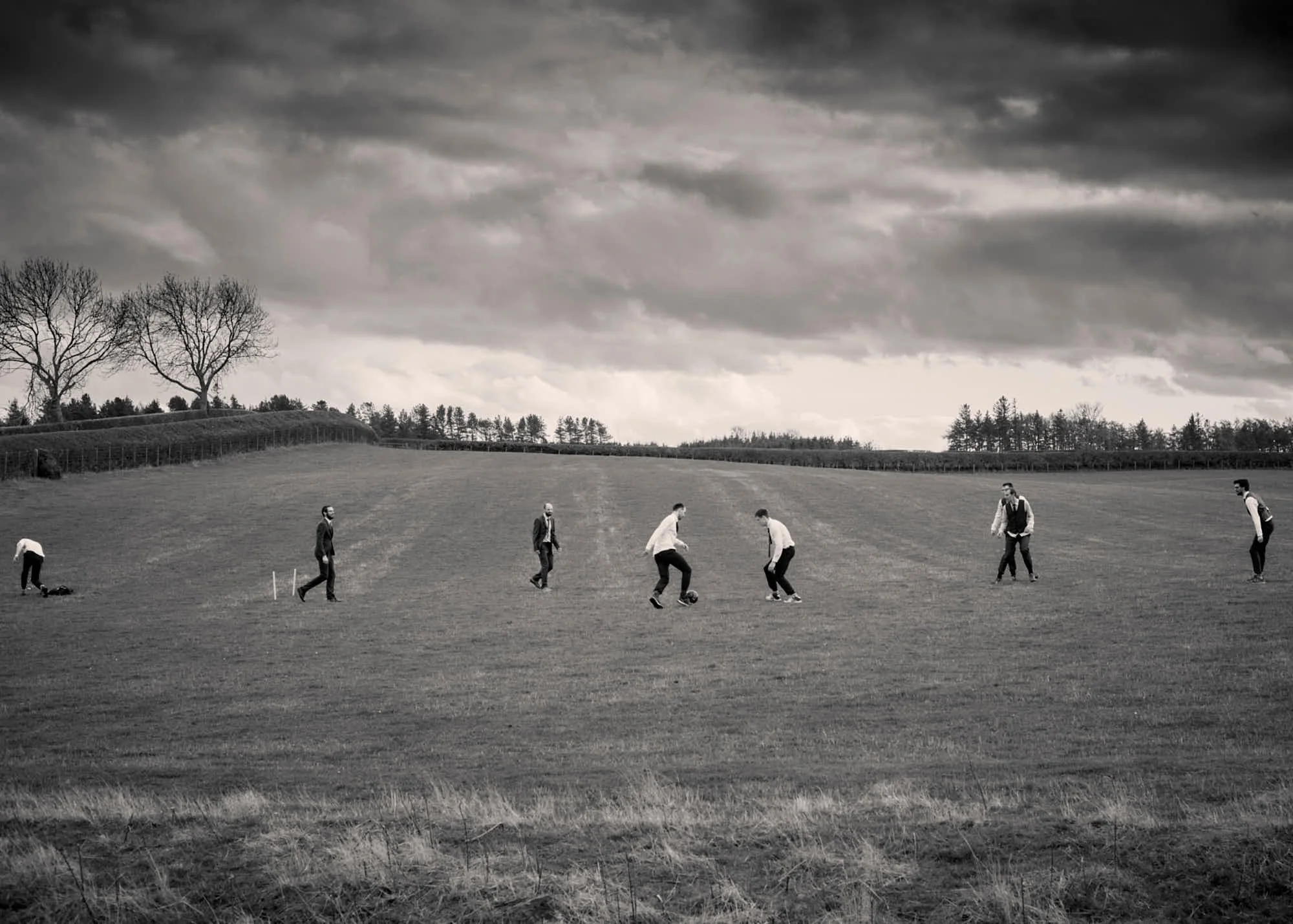 People walking across a grassy field under a dark, cloudy sky with trees and a fence in the background.