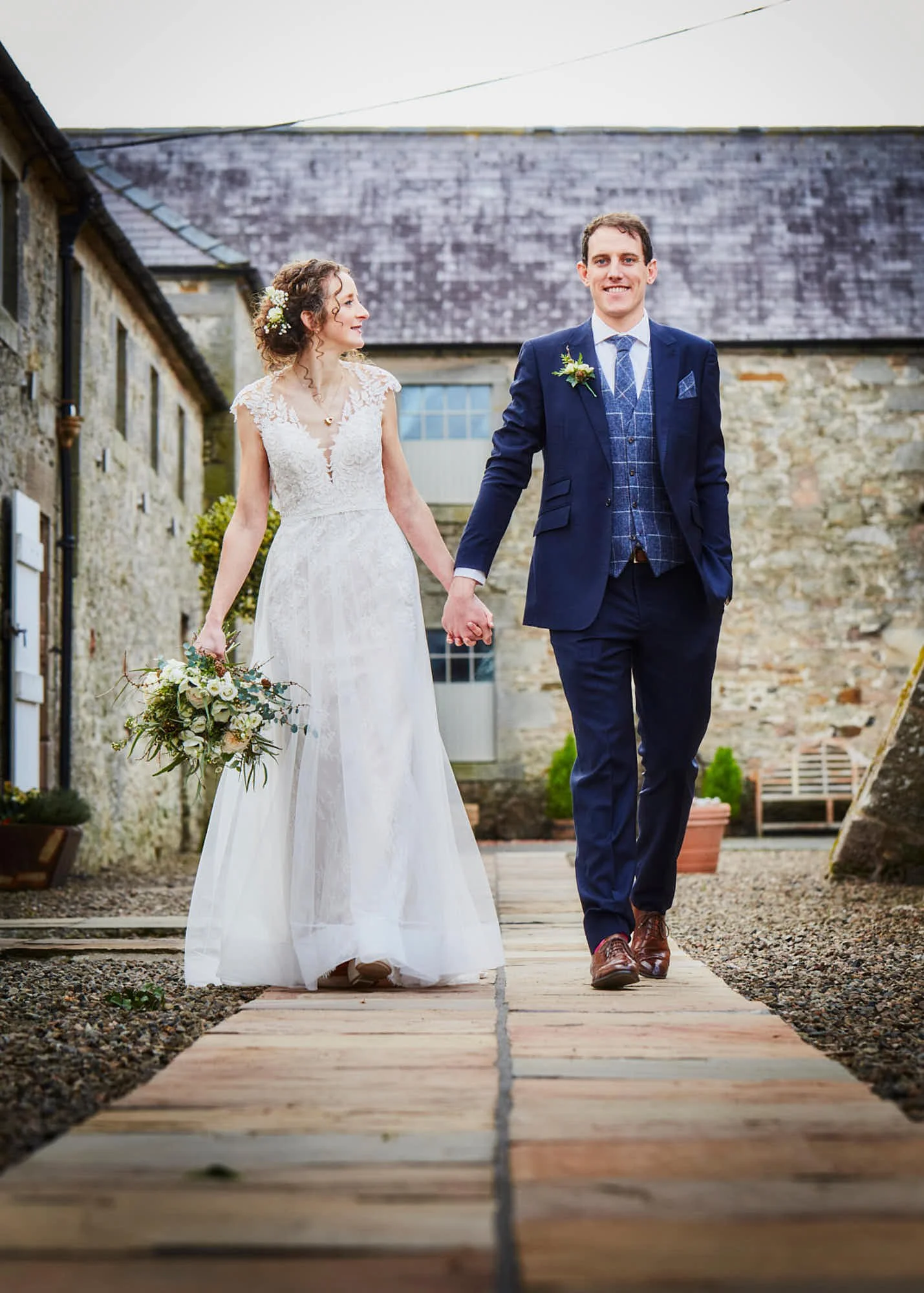 A bride and groom holding hands walking on a wooden pathway outdoors during their wedding, with a rustic stone building in the background.