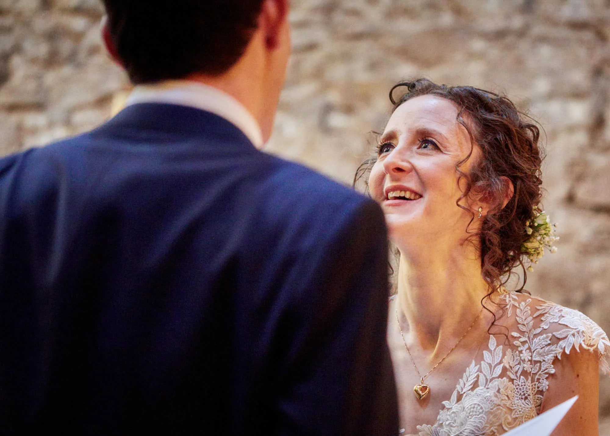 A bride with curly brown hair and a pearl necklace gazes upward at a groom in a dark suit and white shirt during a wedding ceremony.